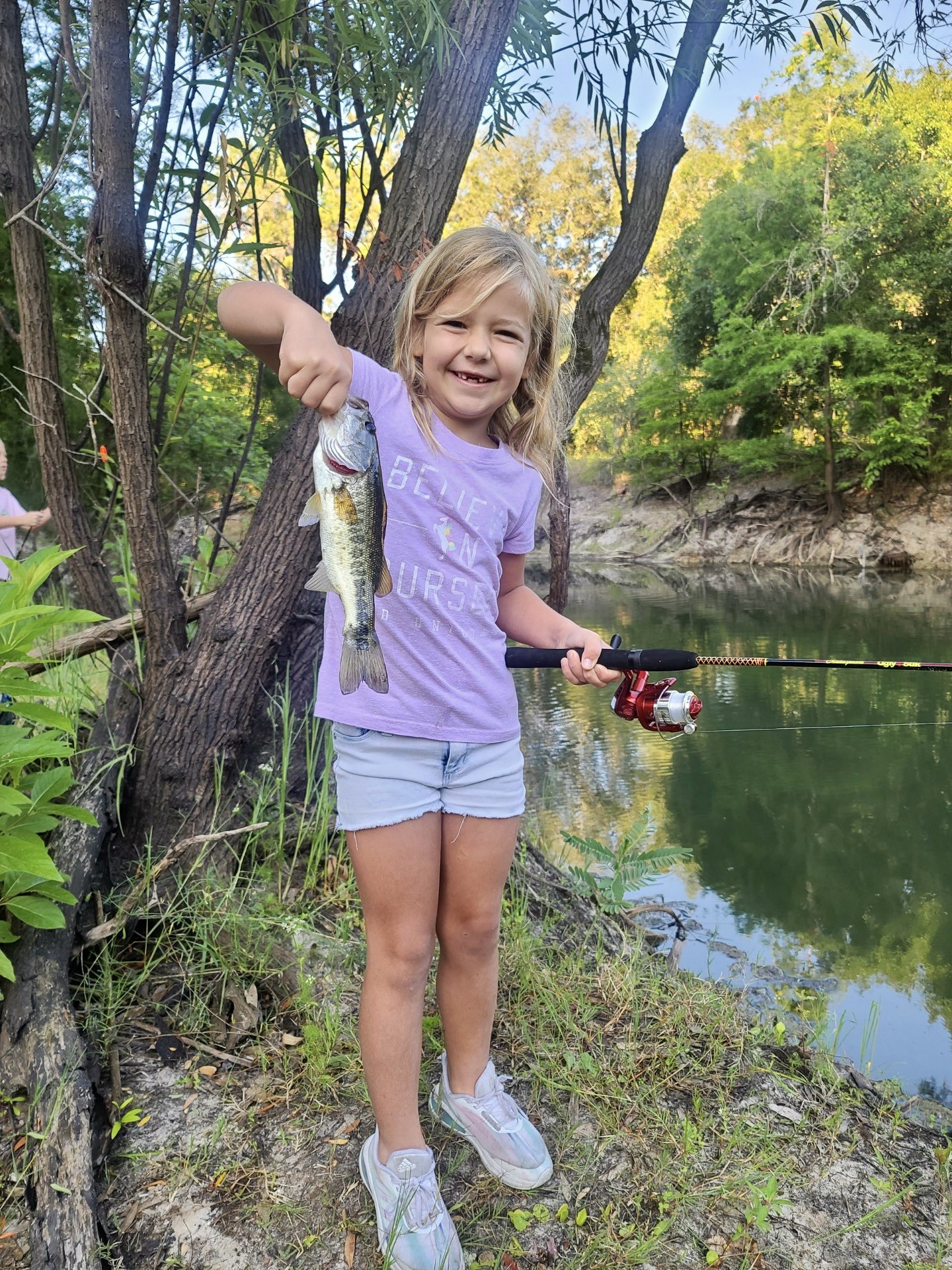 A young girl cheerfully holds up a fish she caught while fishing by a river, with trees and greenery in the background.