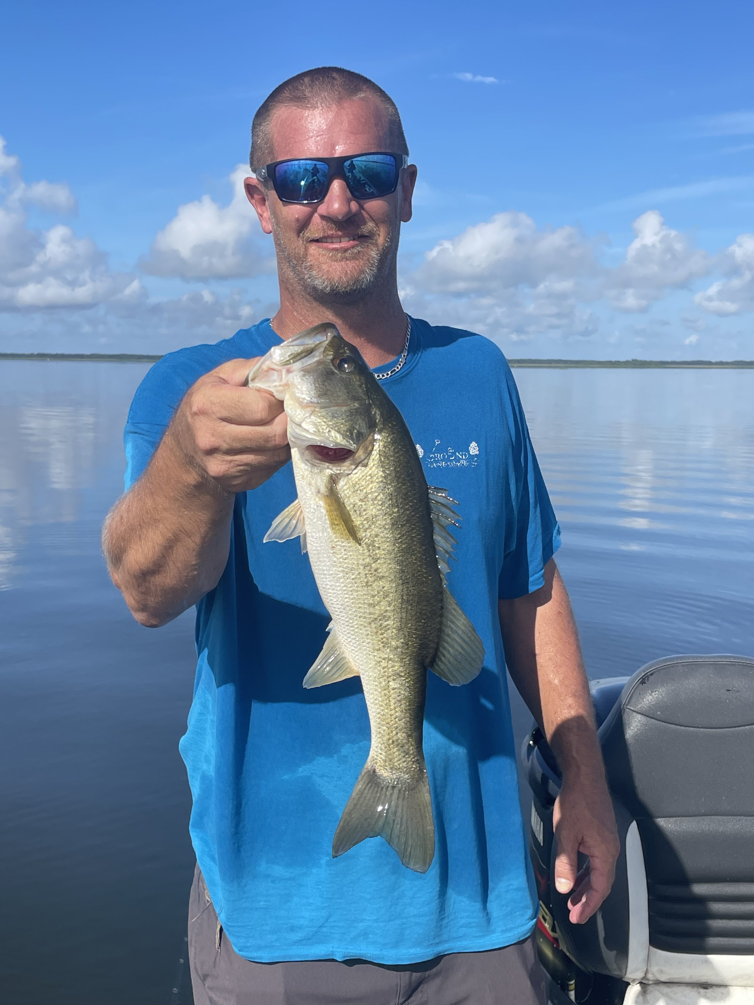 Man wearing sunglasses and a blue shirt holding a largemouth bass fish on a boat with a calm body of water and partly cloudy sky in the background.
