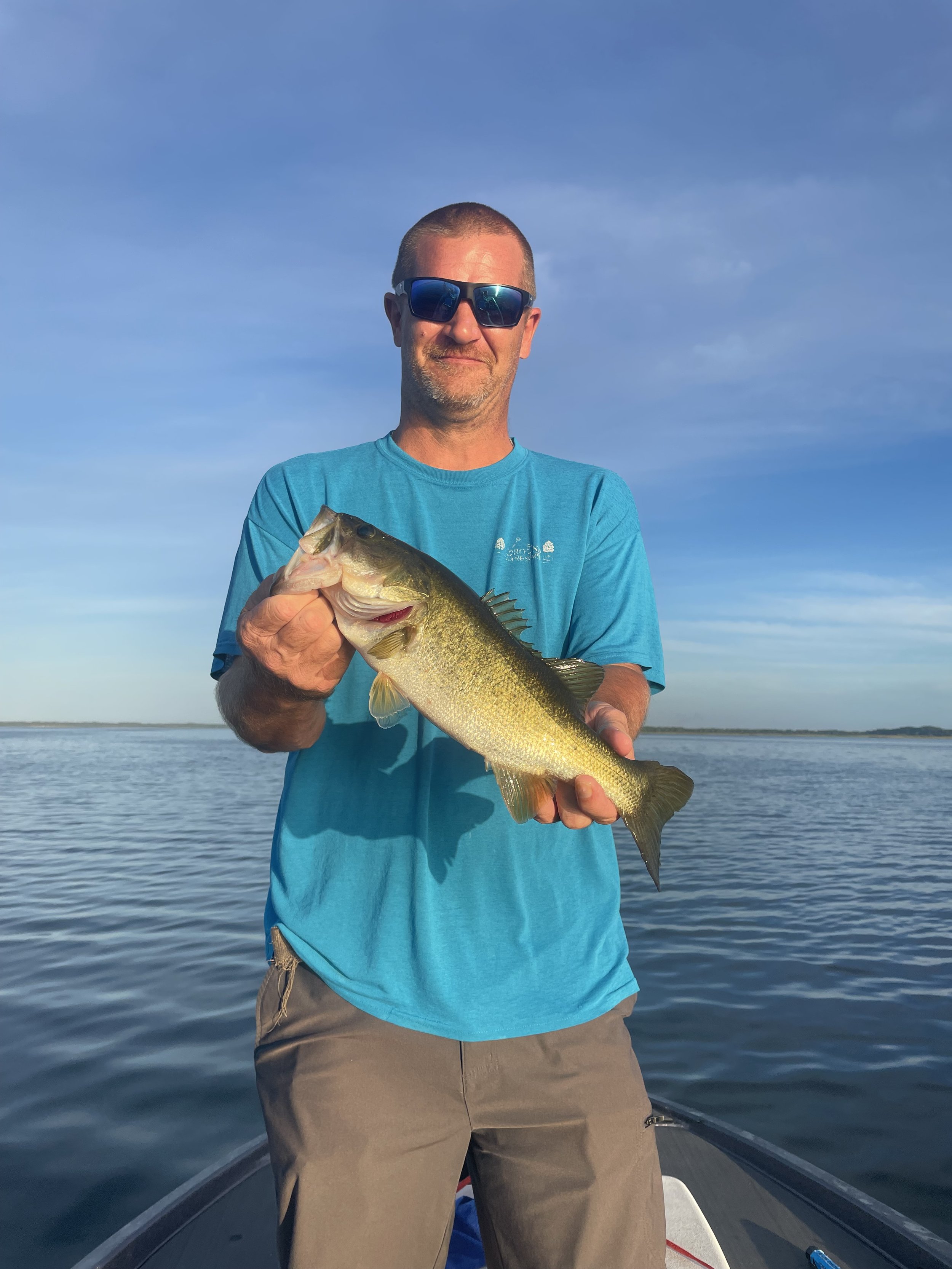 A man wearing sunglasses and a blue T-shirt standing on a boat, holding a large fish, with water and a blue sky in the background.