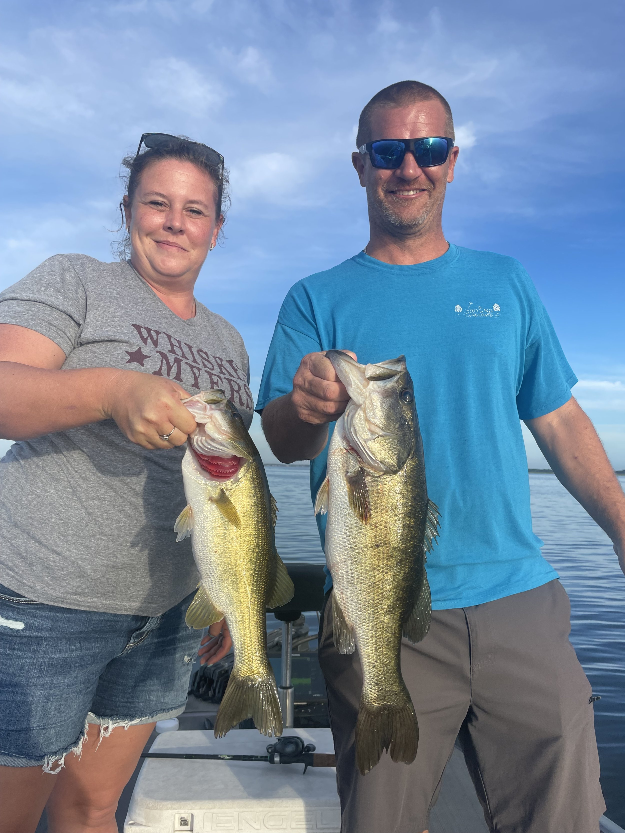 A woman and a man standing on a boat holding two large fish, smiling, with a body of water and a blue sky in the background.