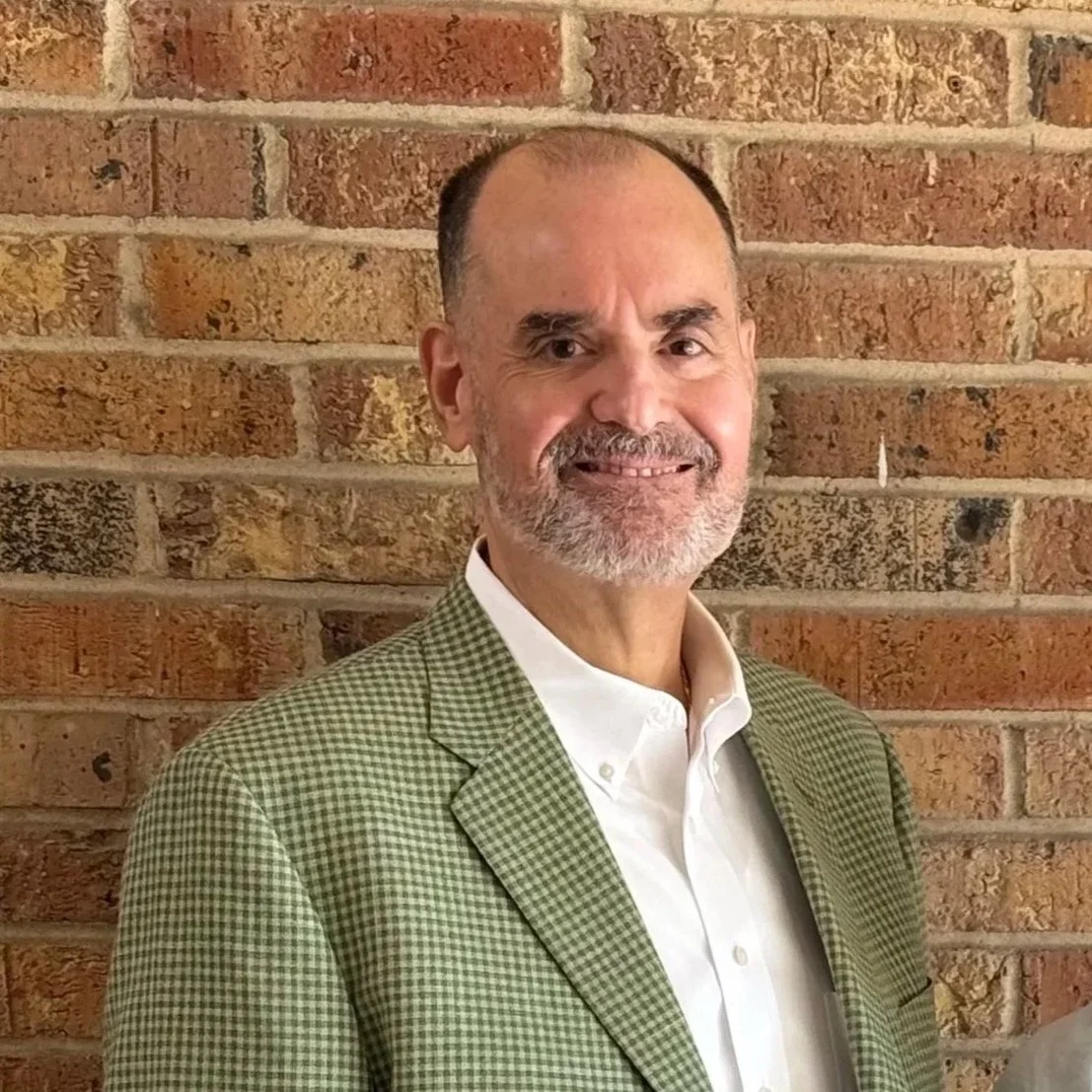 A middle-aged man with a beard and receding hairline smiling, wearing a green checked blazer and white shirt, standing in front of a brick wall.