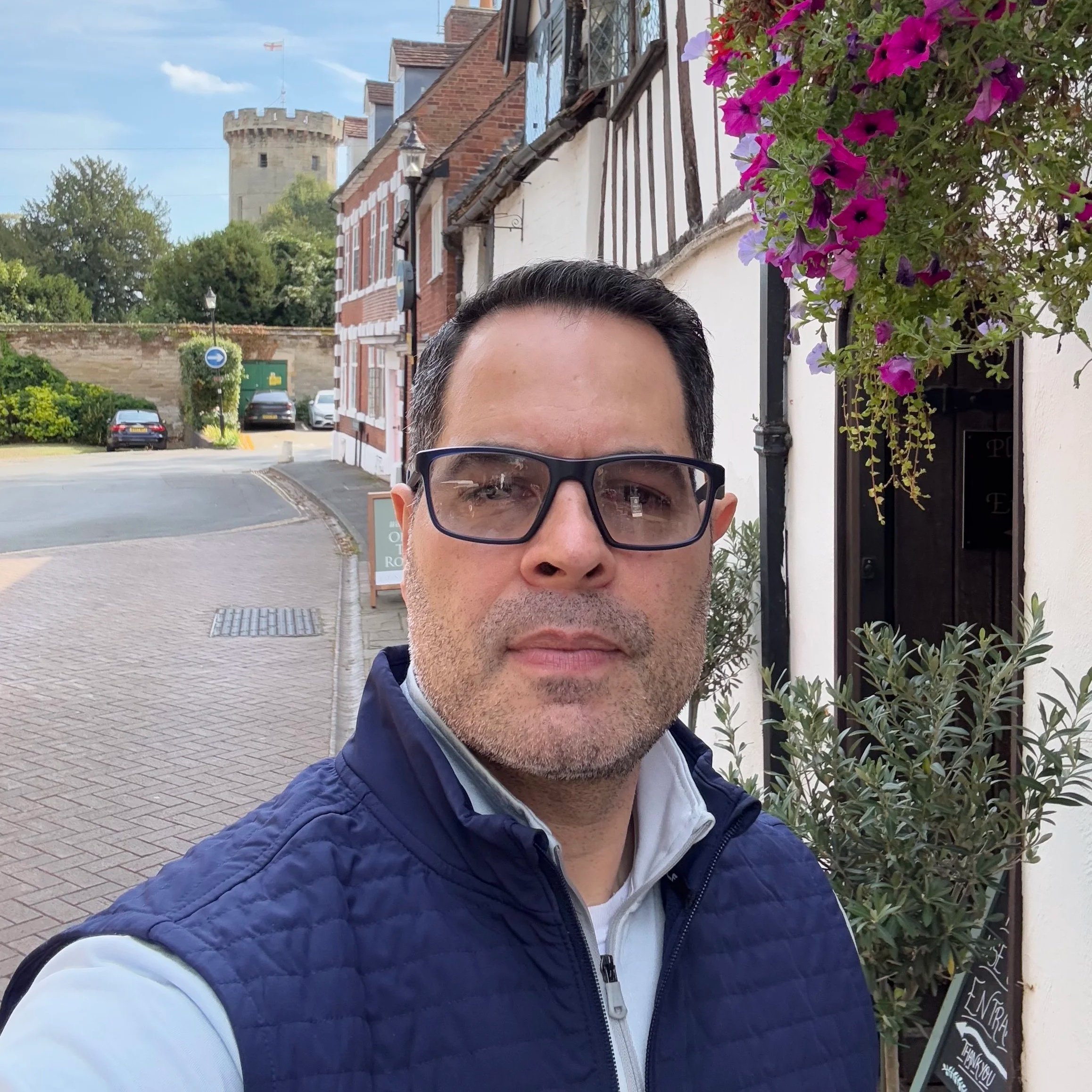 A man with glasses and a blue quilted jacket standing on a cobblestone street next to a white building with flowers and greenery, a tower visible in the background.