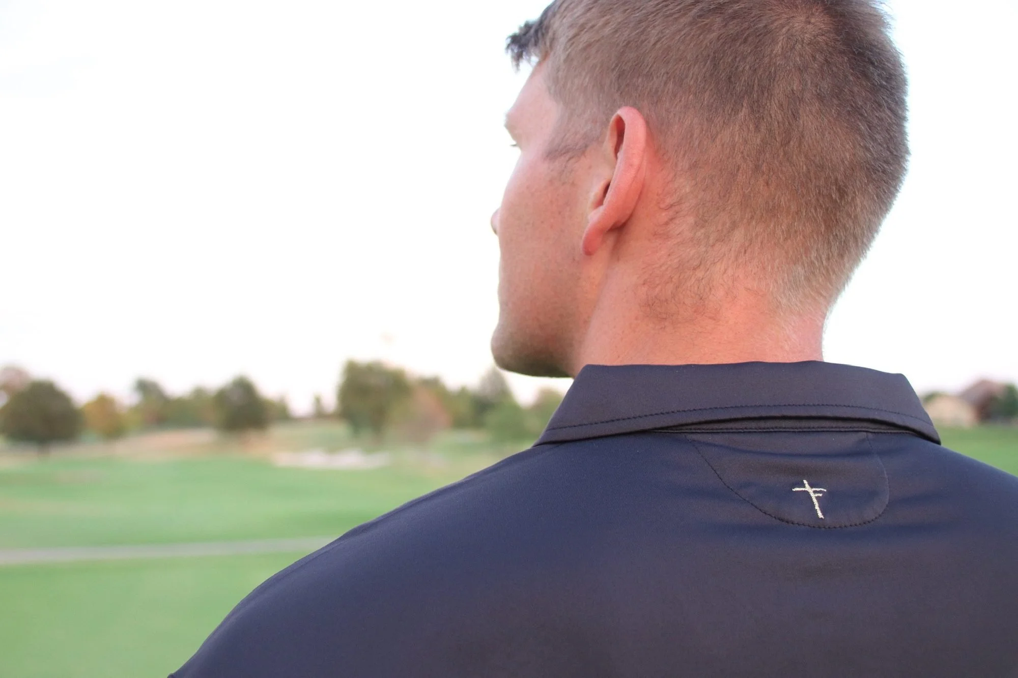 Man on a golf course seen from behind, showing the small embroidered cross logo on the back of a dark Third Fold polo