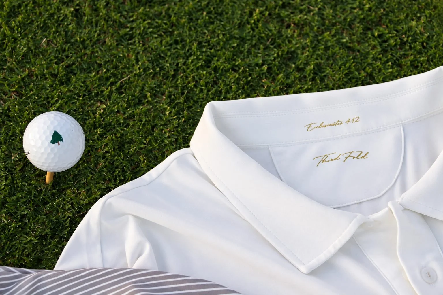 White Third Fold polo on golf course grass beside a teed-up golf ball, with Ecclesiastes 4:12 and Third Fold printed in gold inside the collar