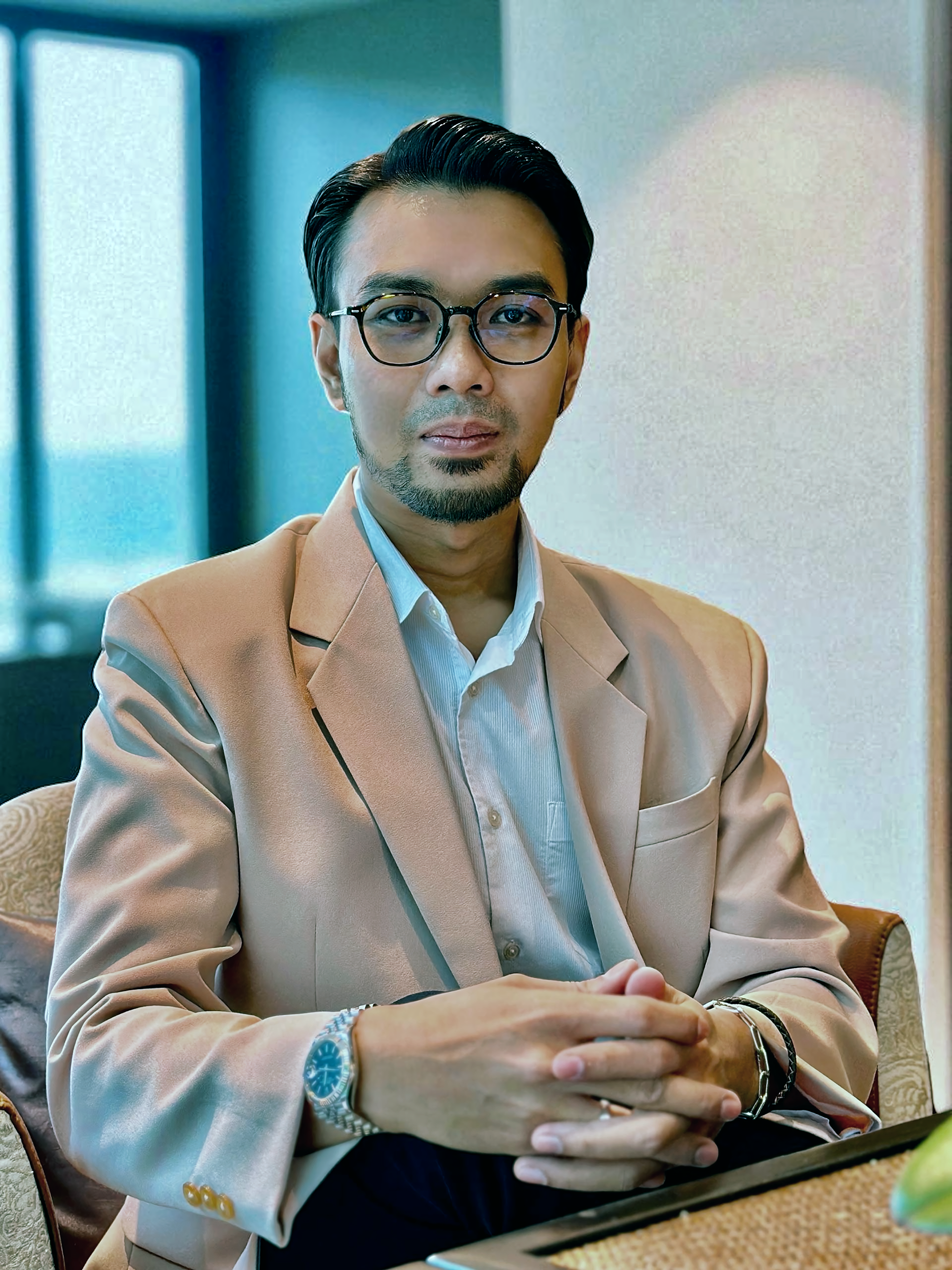 A professional portrait of a man in a black suit, white shirt, black tie, wearing glasses, against a dark background.