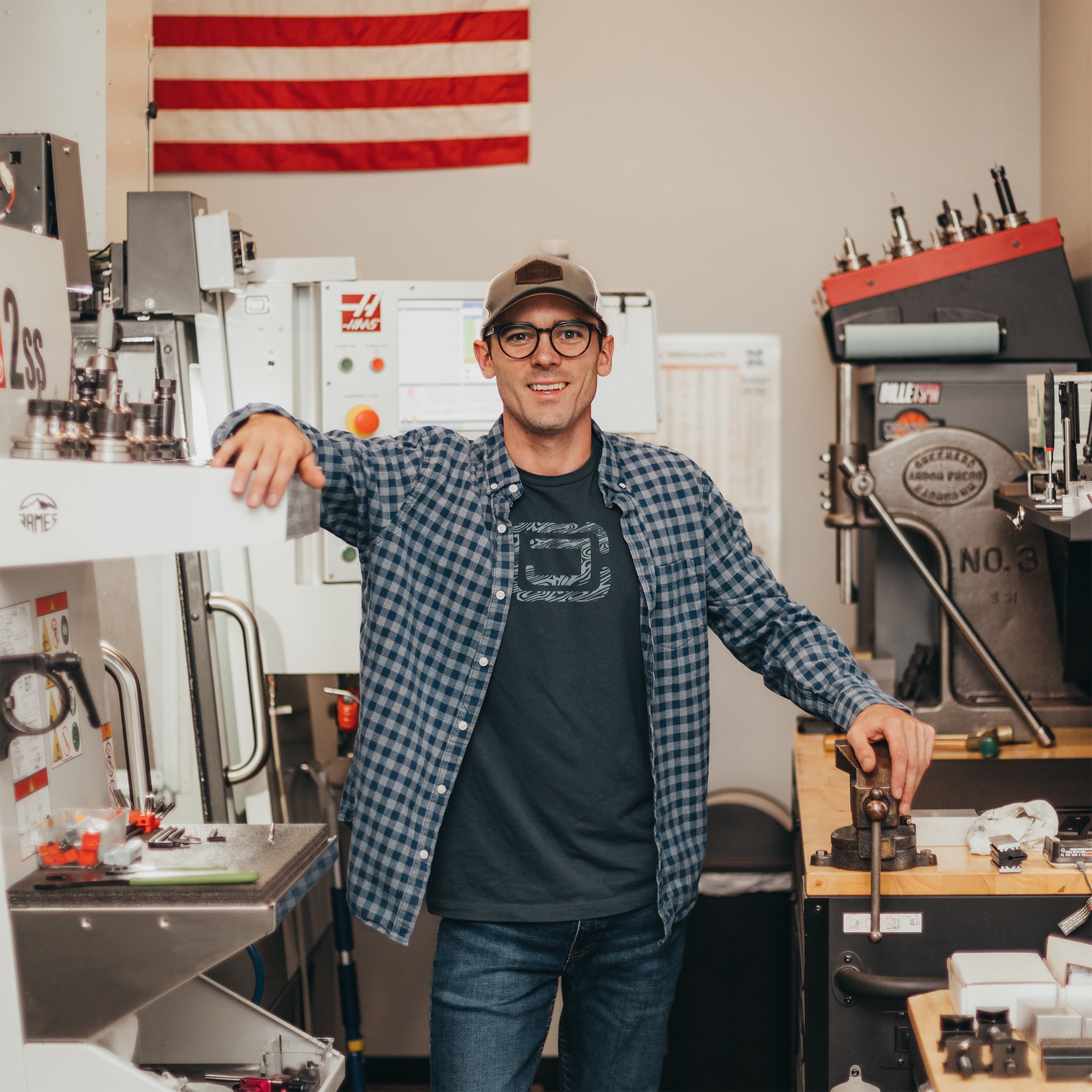 Putter maker Cody James of Cody James Putters standing in his shop beside a CNC machine with an American Flag hanging in the background.