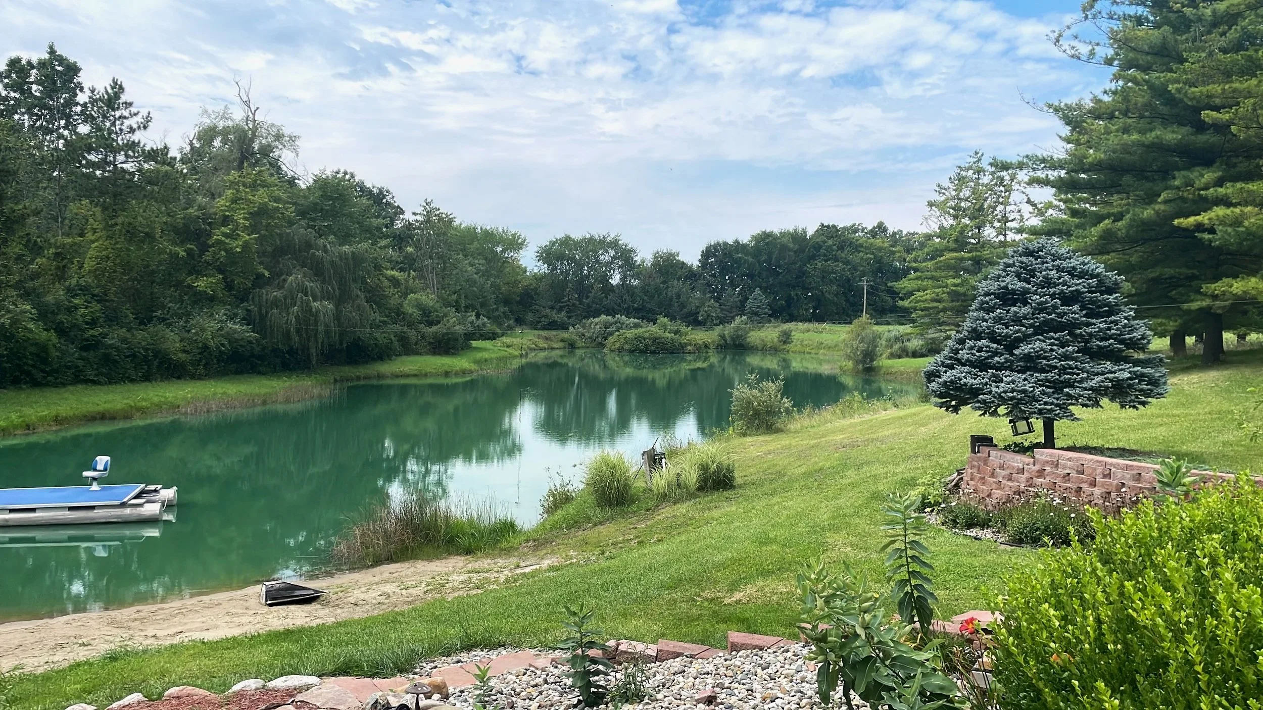 A peaceful lakeside scene with green trees, a small boat on the water, and a manicured lawn with a decorative blue-green tree and a brick border garden.