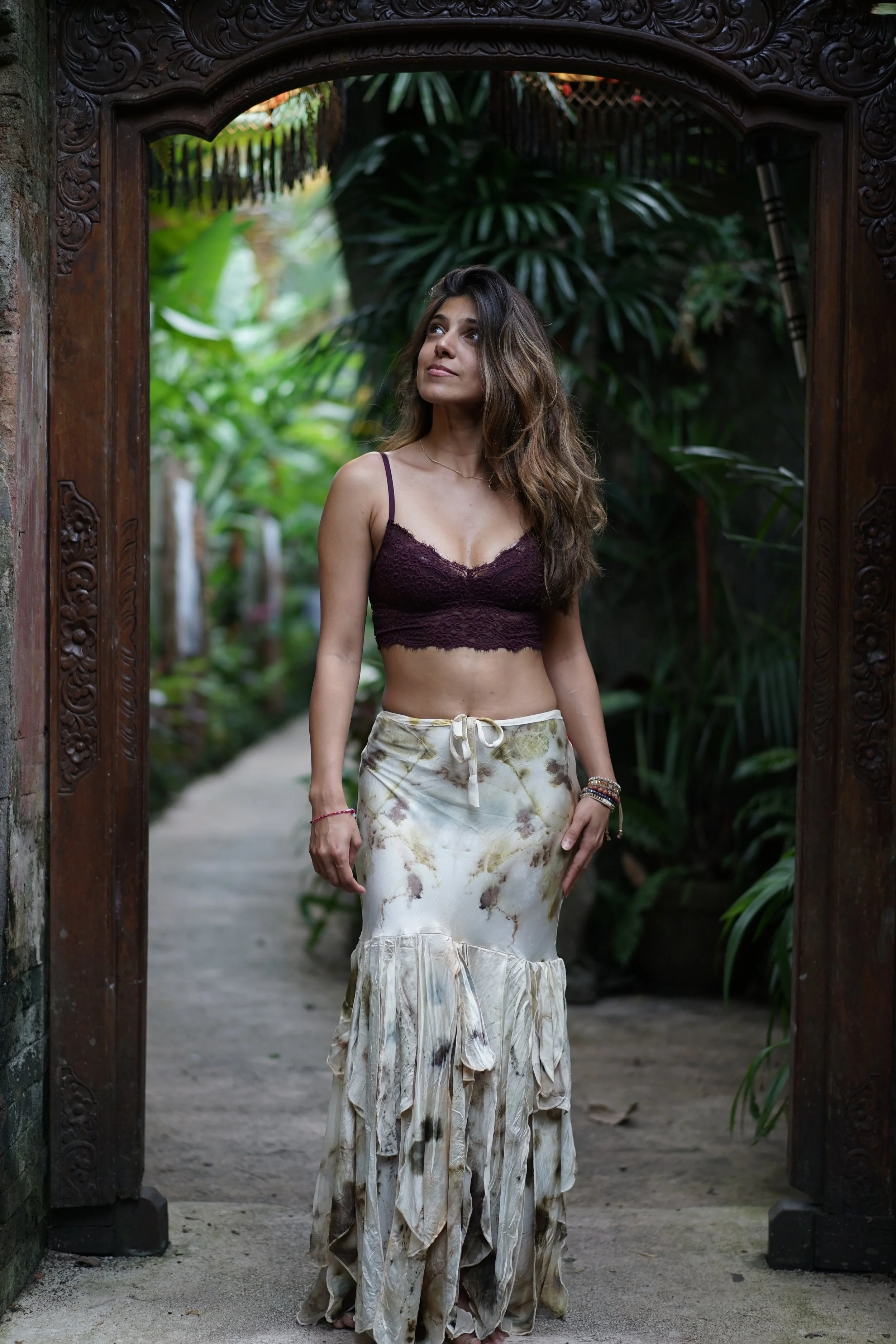 A young woman standing in a garden doorway, wearing a burgundy lace crop top and a long, flowy skirt with a floral pattern.