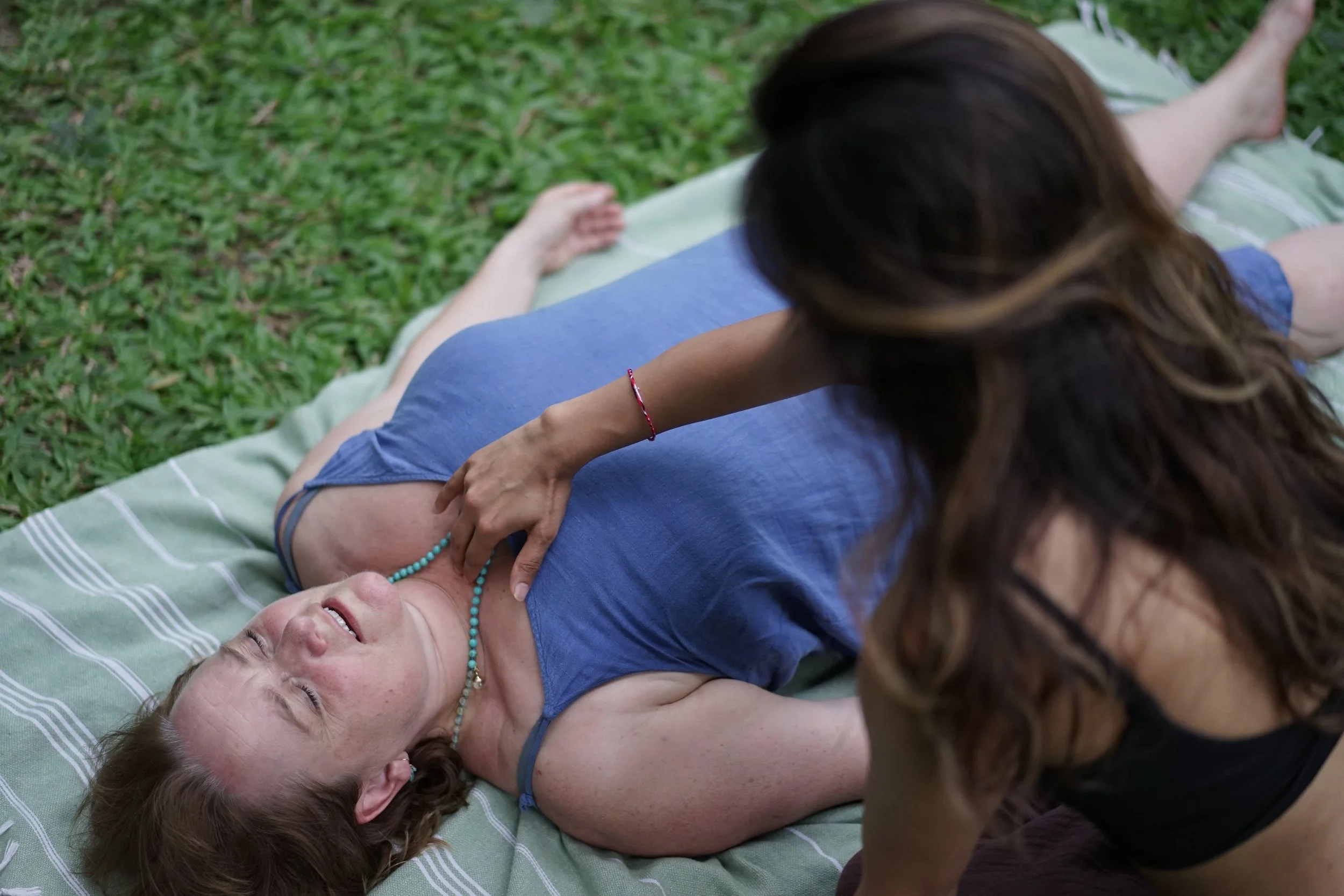 A woman lies on a blanket outdoors on grass, appearing to be in a relaxed or happy state, while another woman leans over her and touches her chest.