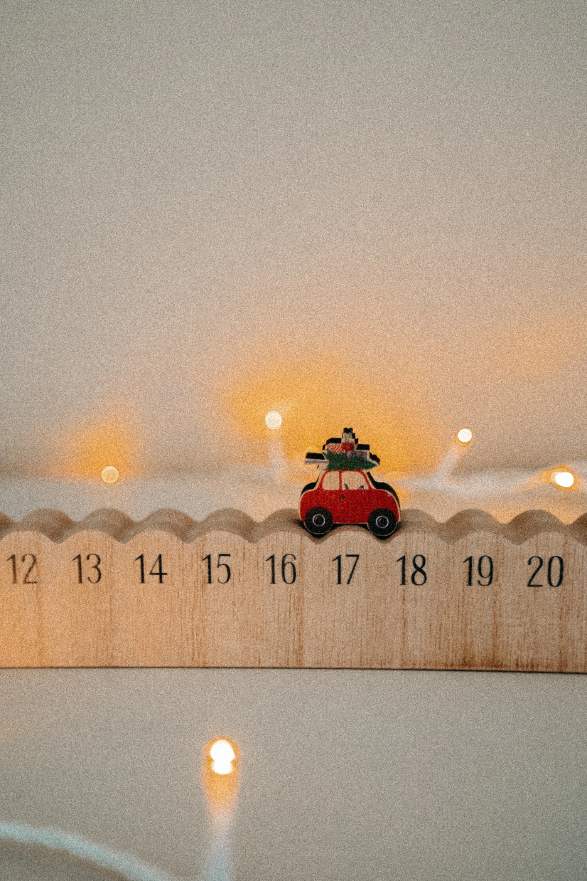 Puinen adventtikalenteri, jonka päällä on pieni auto- ja joululahjasetin, ja ympärillä lämpöisiä valoja.