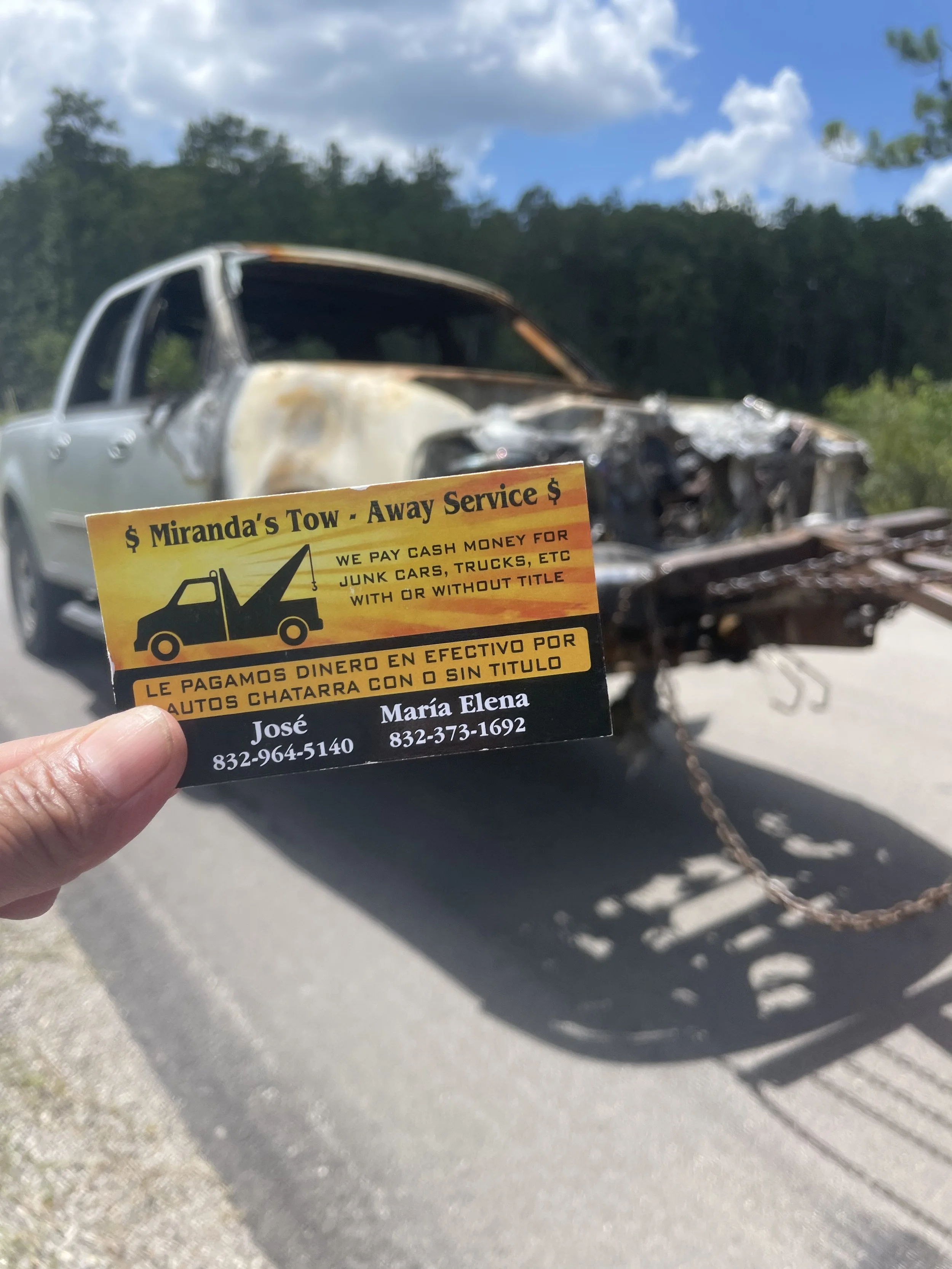 Hand holding a business card for Miranda's Tow in front of a burnt-out, wrecked vehicle on the side of the road, with a background of trees and blue sky.