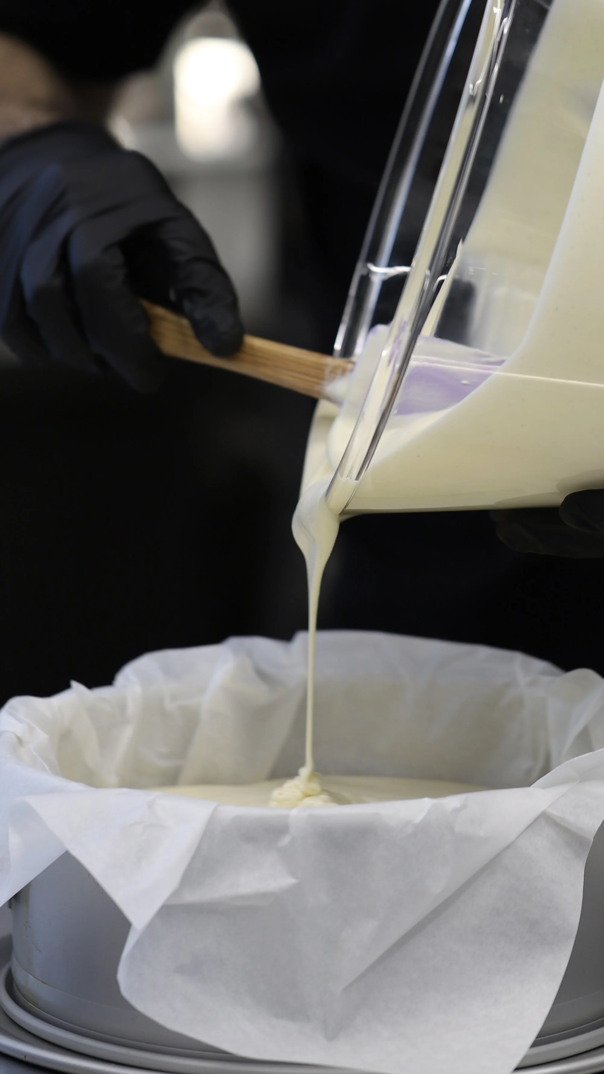 A person wearing black gloves is pouring liquid cheese into a cheese-making mold lined with cheesecloth.