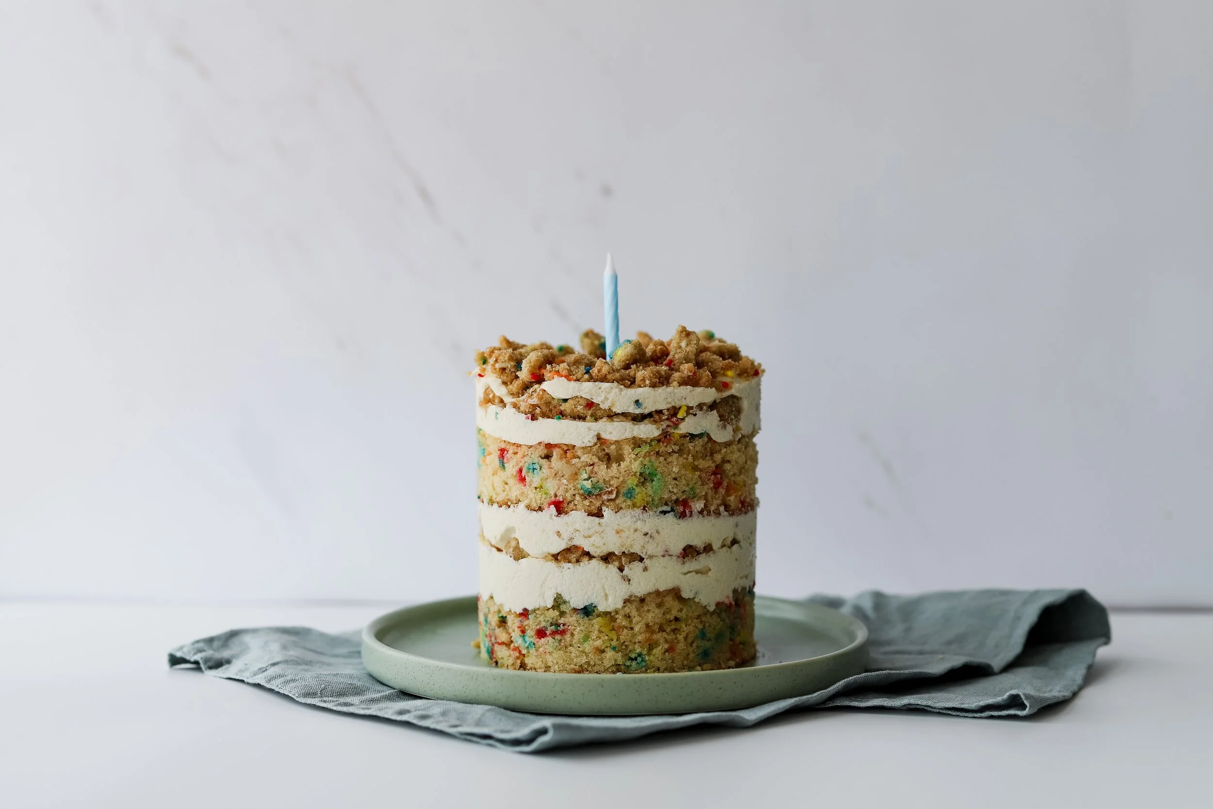 A tower of cookies and cream ice cream sandwiches with colorful cookie crumbs and sprinkles, topped with a birthday candle, on a white plate with a gray napkin underneath.