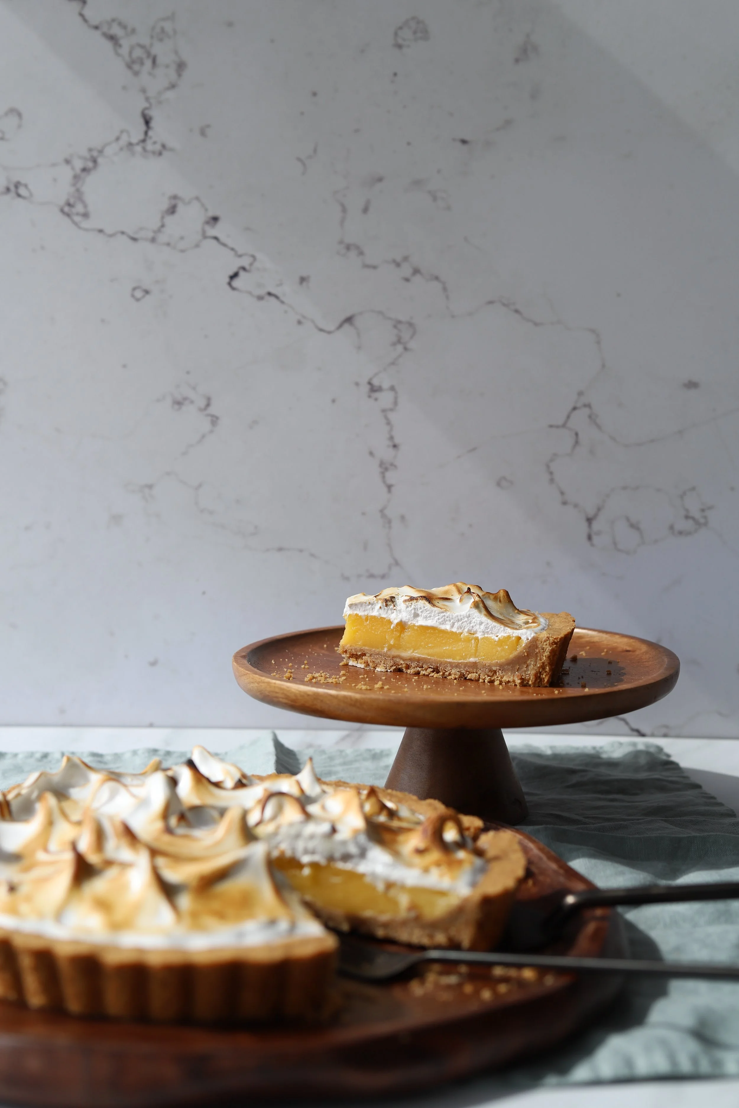 Close-up of a lemon meringue pie with toasted meringue peaks on a wooden serving plate, with a slice cut out revealing lemon filling, on a marble surface with another pie in the background against a white marble wall.