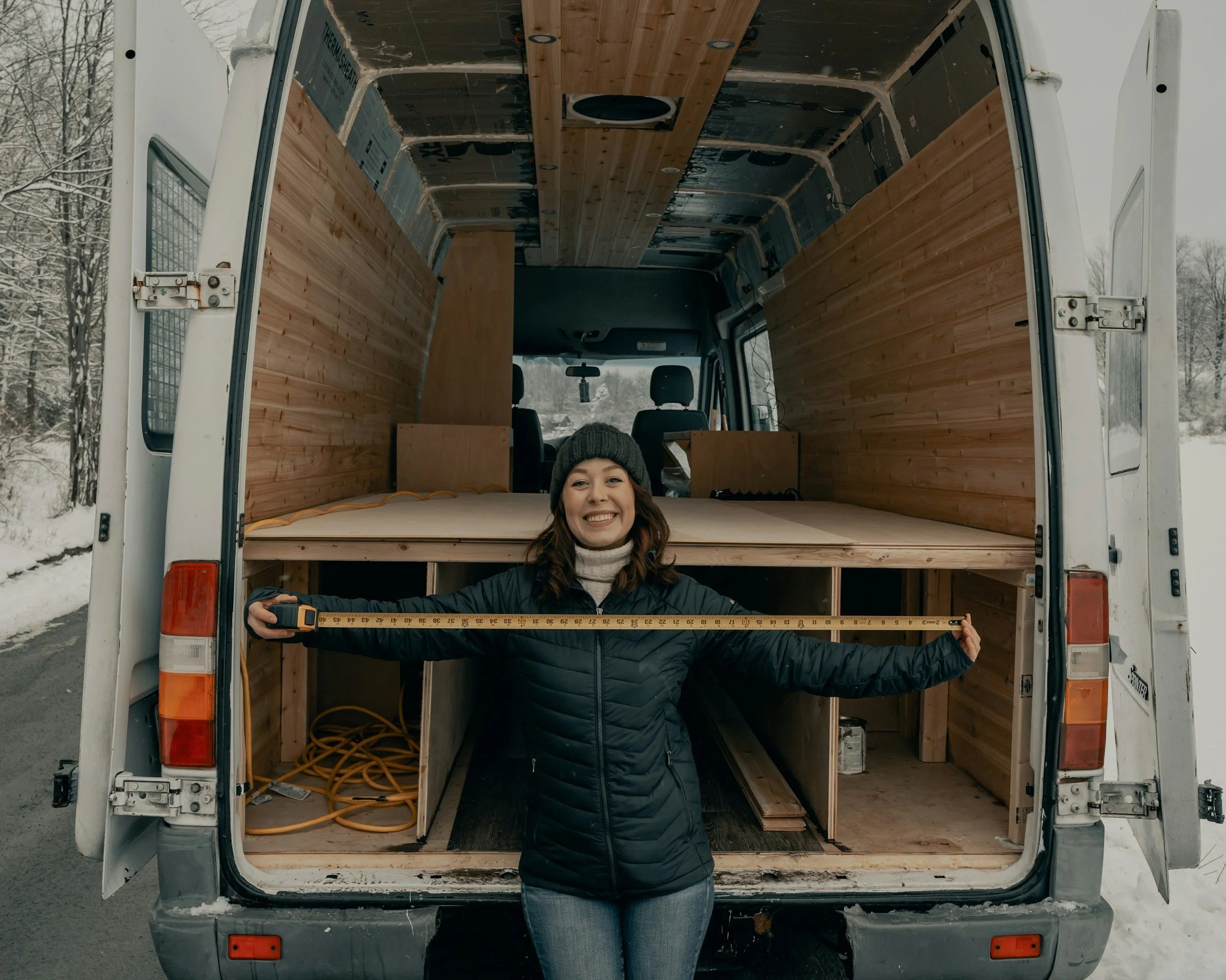 A woman smiling and holding a measuring tape in front of an empty wood-paneled van, with an outdoor snowy landscape in the background.