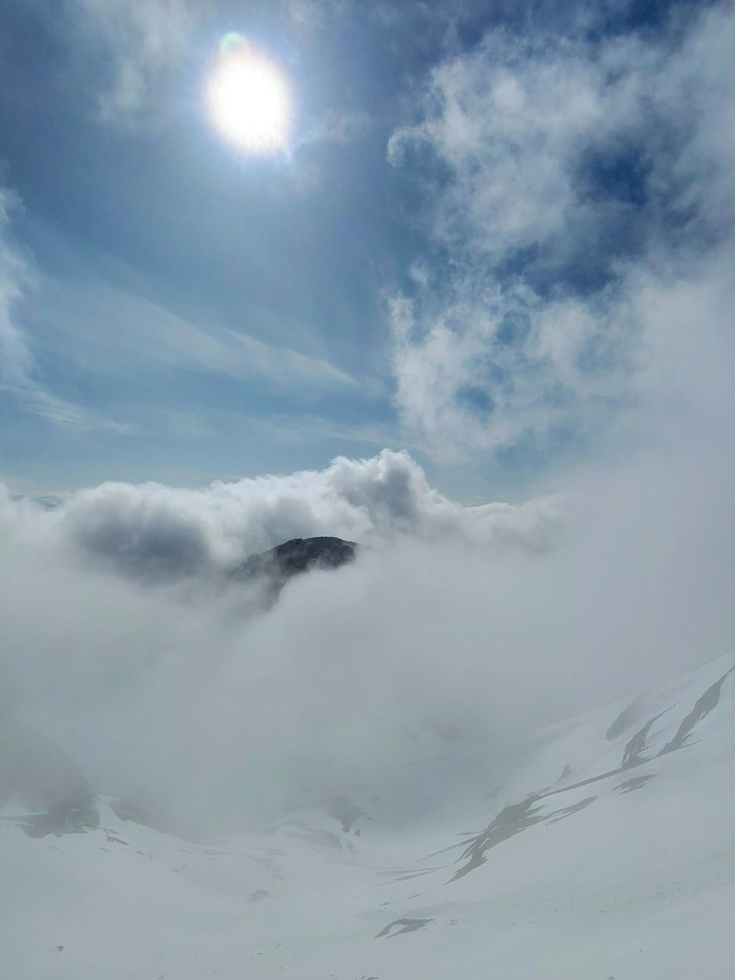 A snow-covered mountain top surrounded by clouds and visible through a window from inside an aircraft, with the sun shining in the clear blue sky.