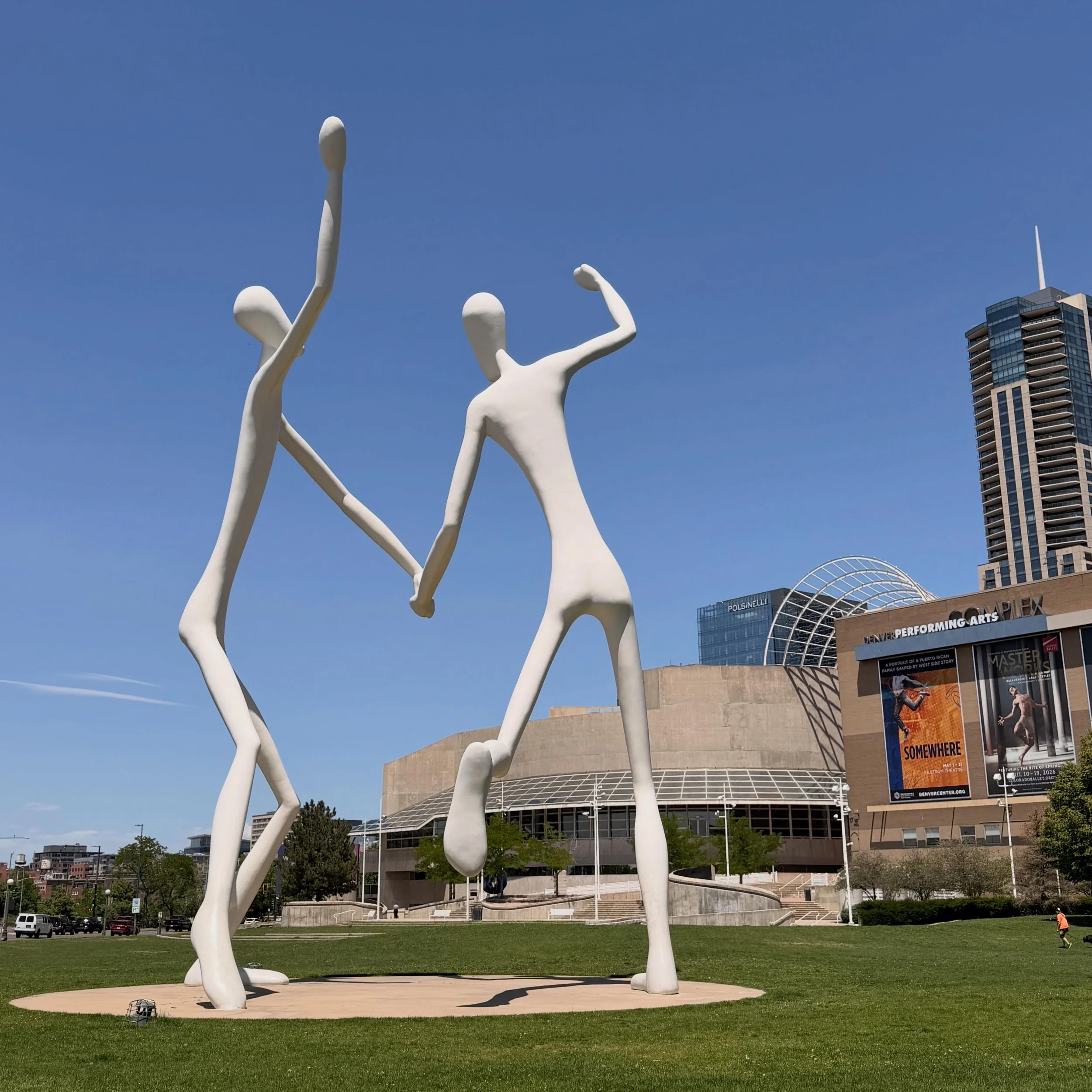 Sculpture of abstract human figures made of white material, located in an outdoor urban park with grass and buildings in the background.