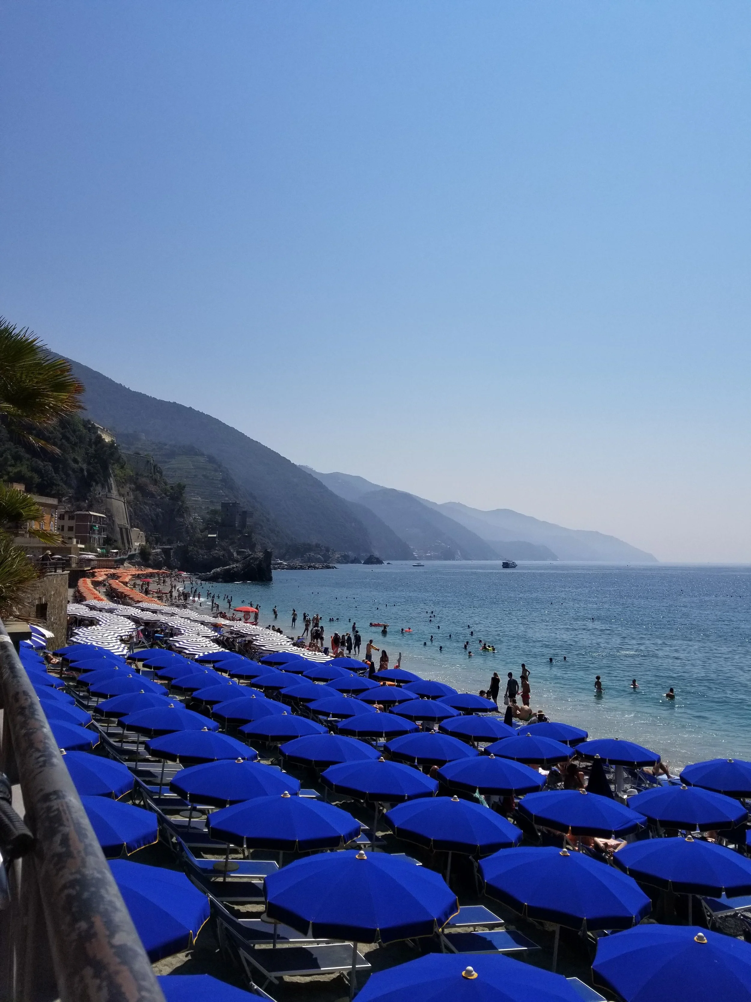 Beach scene with blue sun umbrellas, people swimming and relaxing in the ocean, hillside with buildings and greenery in the background, and a clear blue sky.