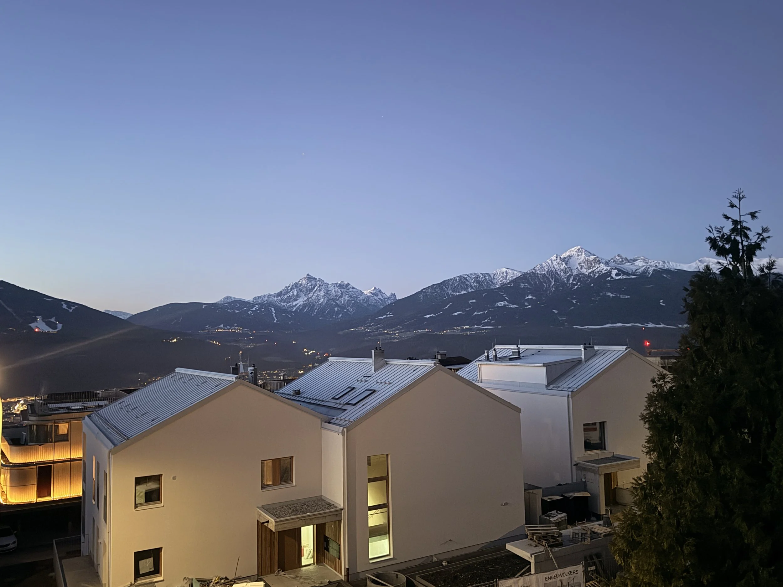 Nighttime mountain scene with snow-capped peaks and a partly cloudy sky, viewed from a residential area with modern buildings and a tree on the right.