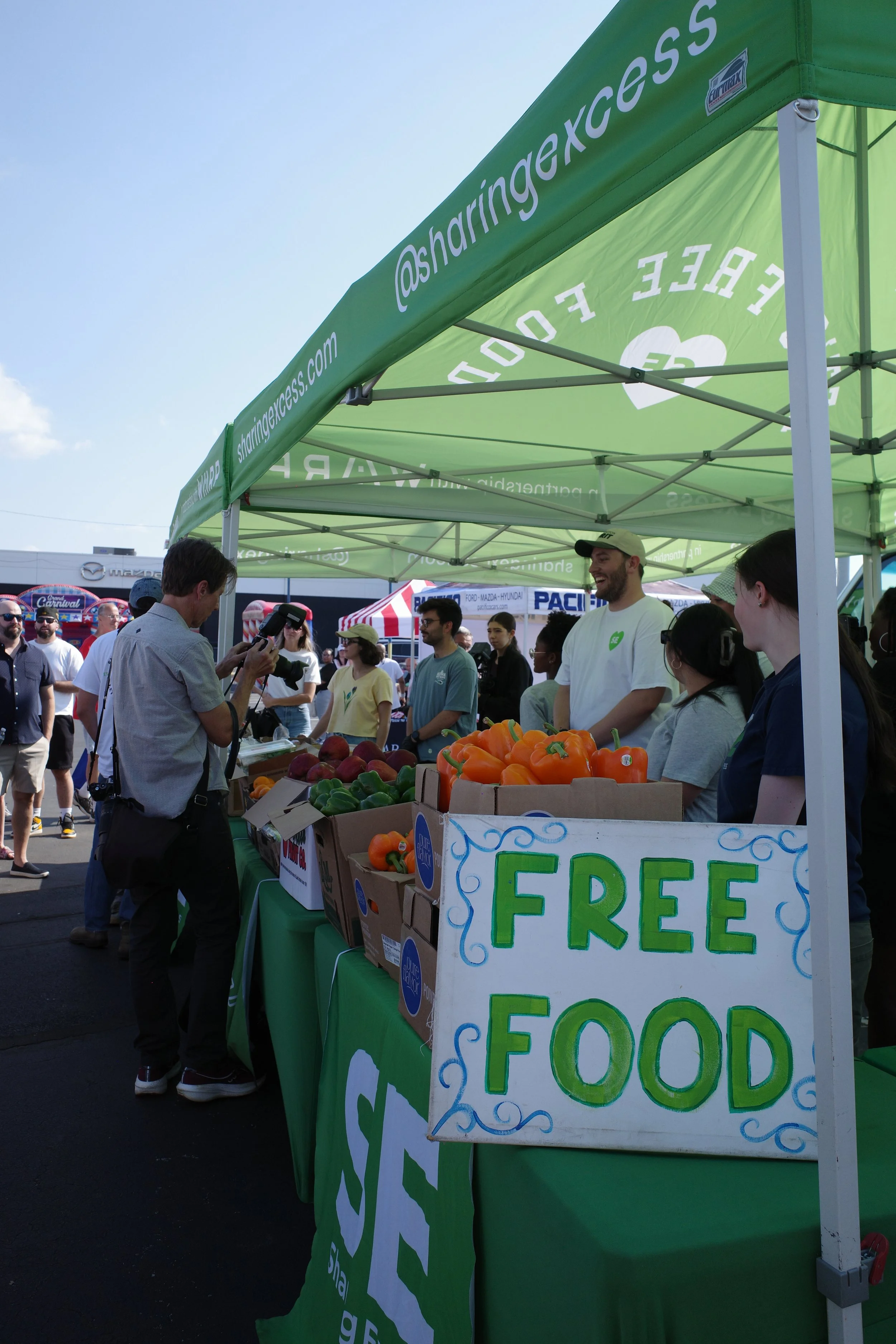 People at a market stand with a sign that reads 'Free Food' and a canopy with the social media handle '@sharingxcess'. There are fresh vegetables on the table and several people shopping or talking.