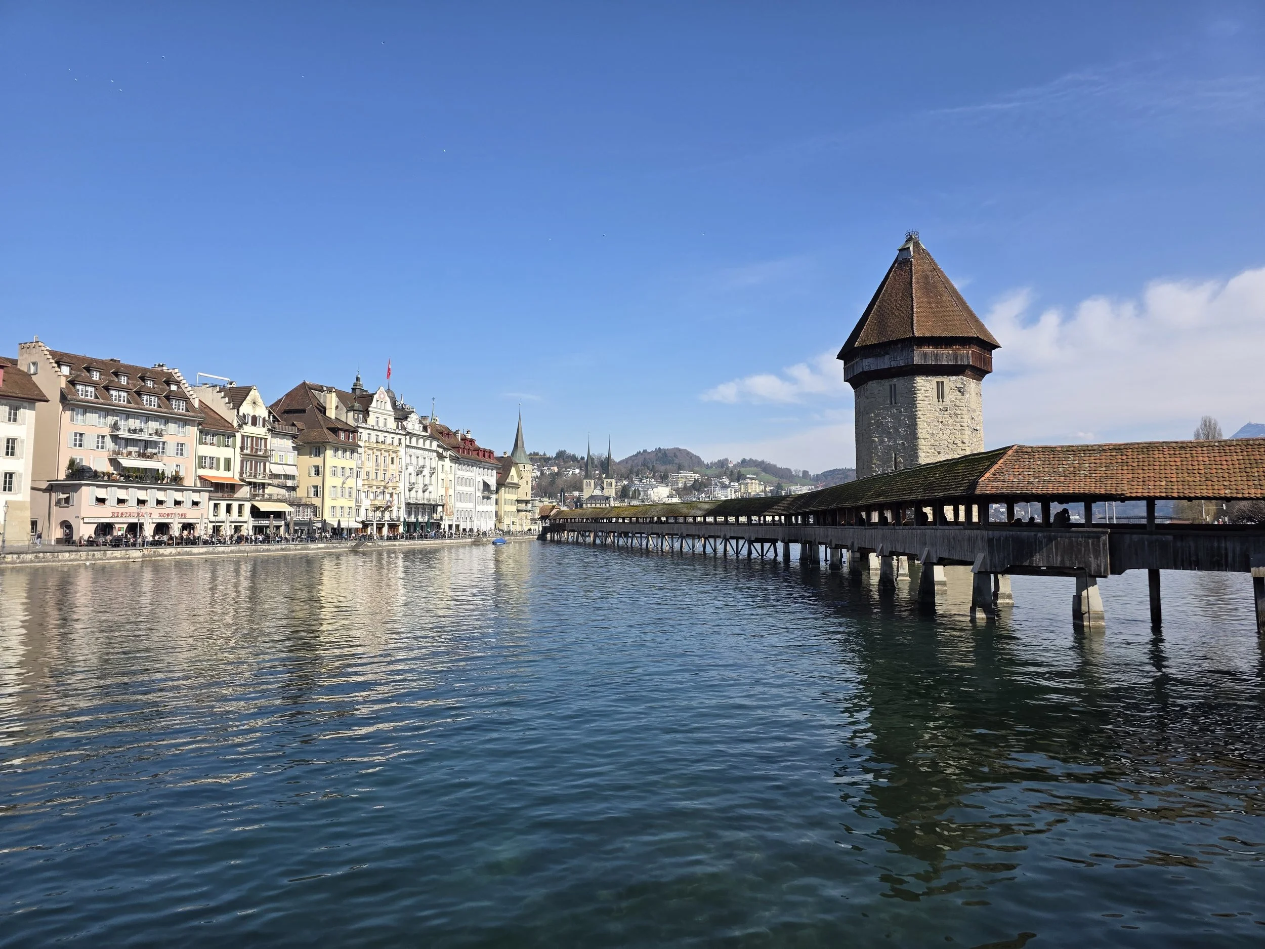 A cityscape with colorful buildings along a river, featuring a large medieval stone tower with a wooden roof. The sky is mostly clear with a few clouds and the water reflects the buildings and sky.