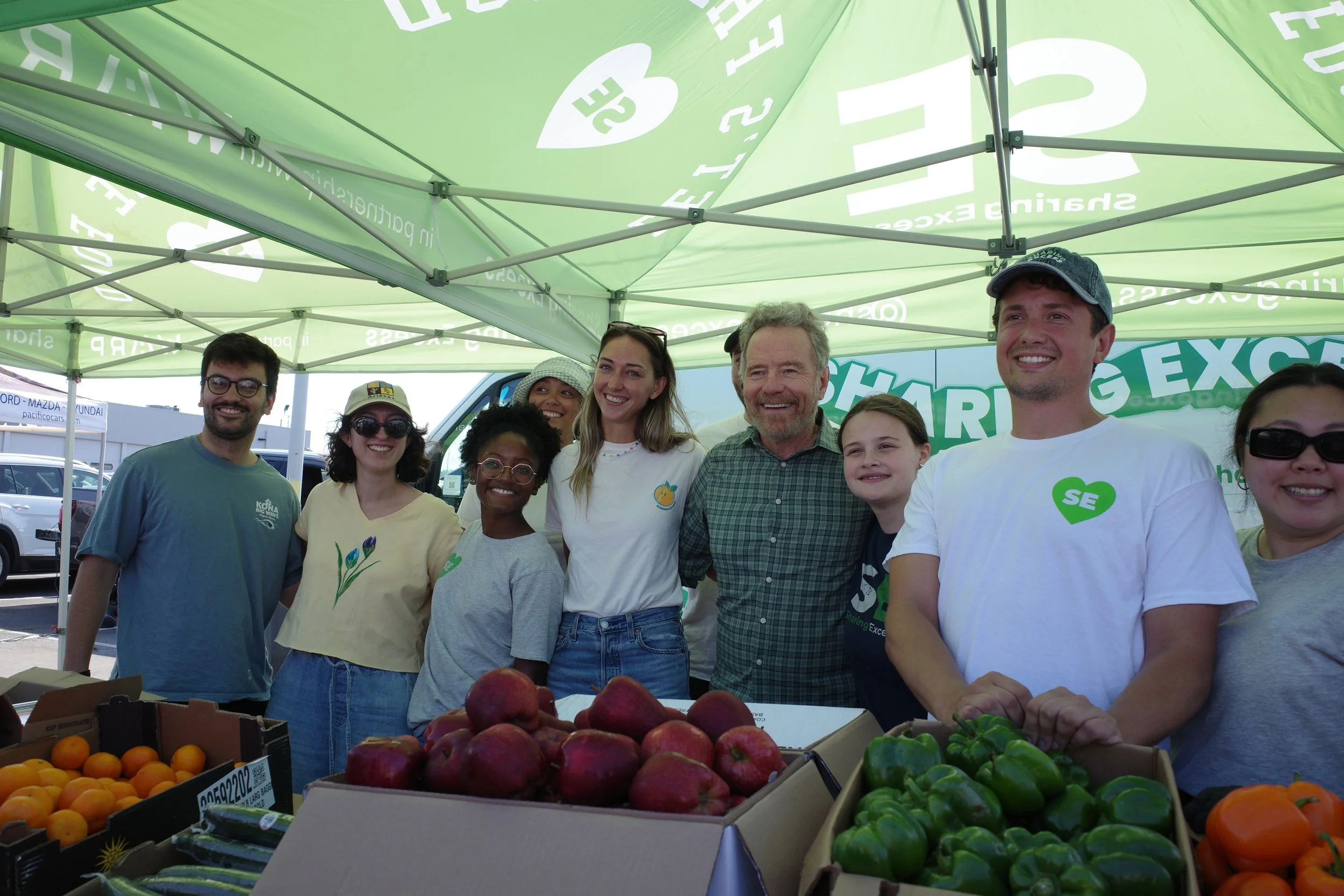 A diverse group of people standing under a green canopy at a farmers market, with fresh fruits and vegetables like peaches, green peppers, and oranges in front of them.