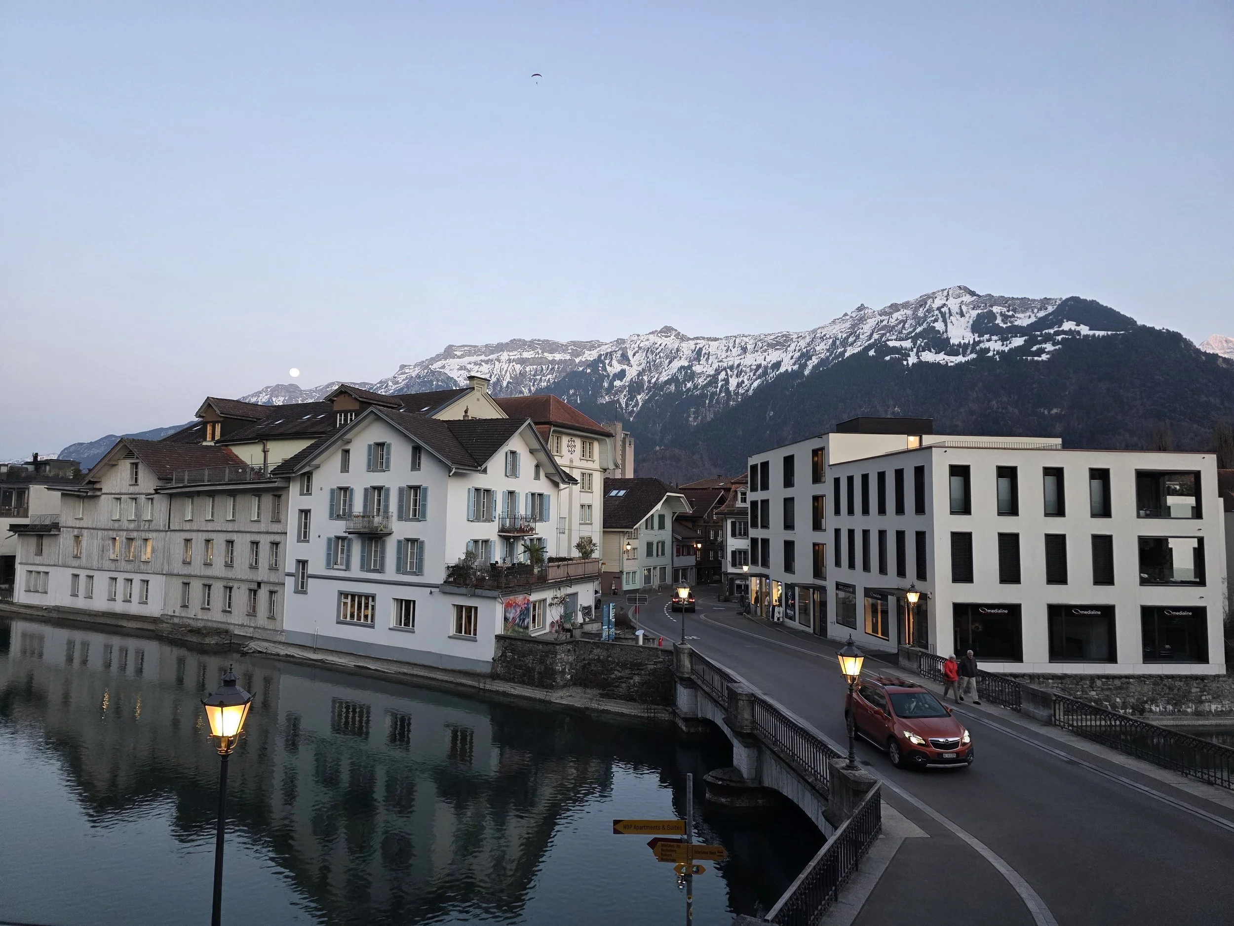 A scenic view of residential buildings by a river with snow-capped mountains in the background during dusk or early evening.