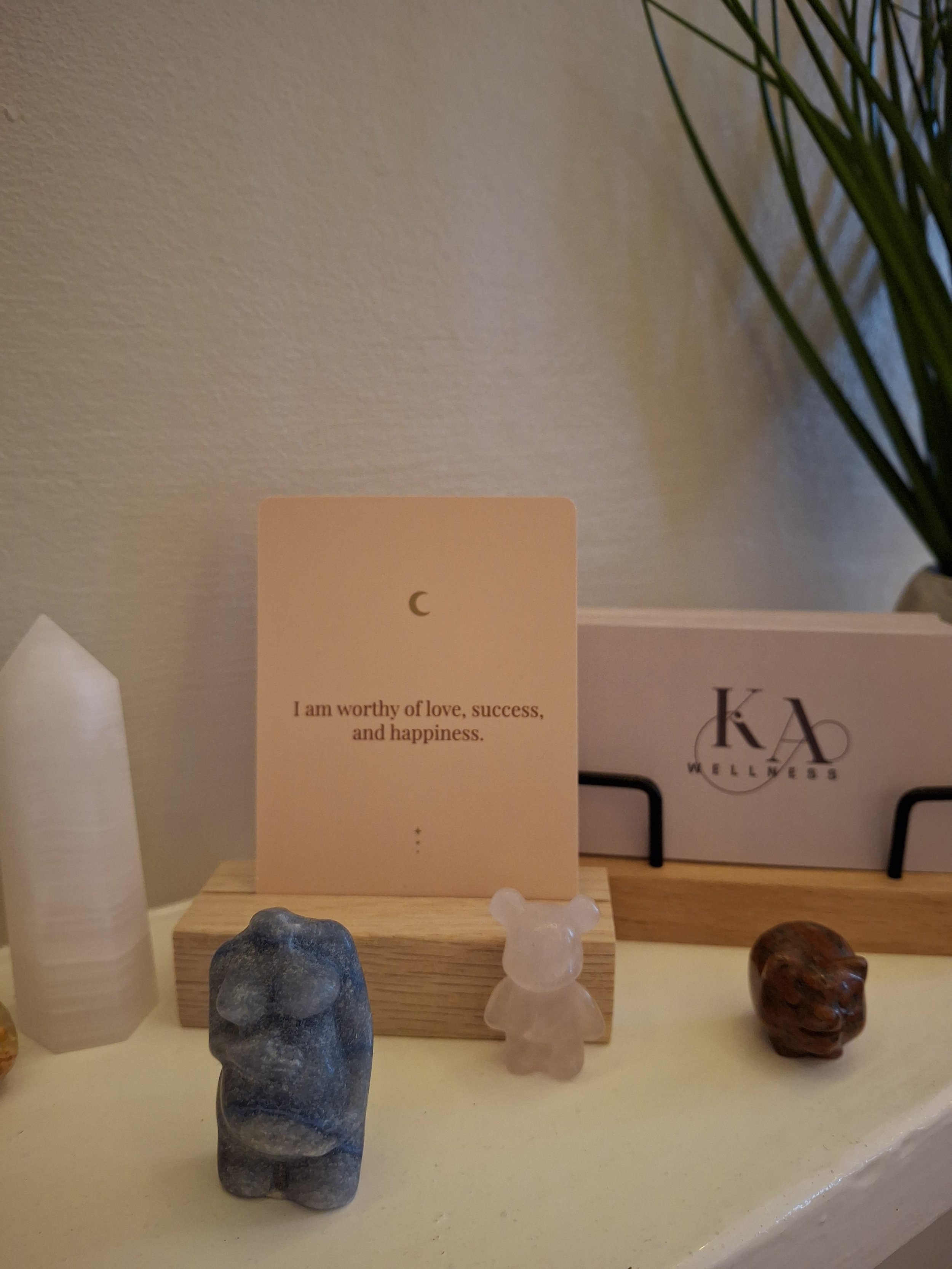 A small white card with a crescent moon symbol and the words "I am worthy of love, success, and happiness" placed on a wooden stand. Surrounding it are small decorative stones, including a blue stone, a pinkish bear-shaped stone, and a brown stone, o