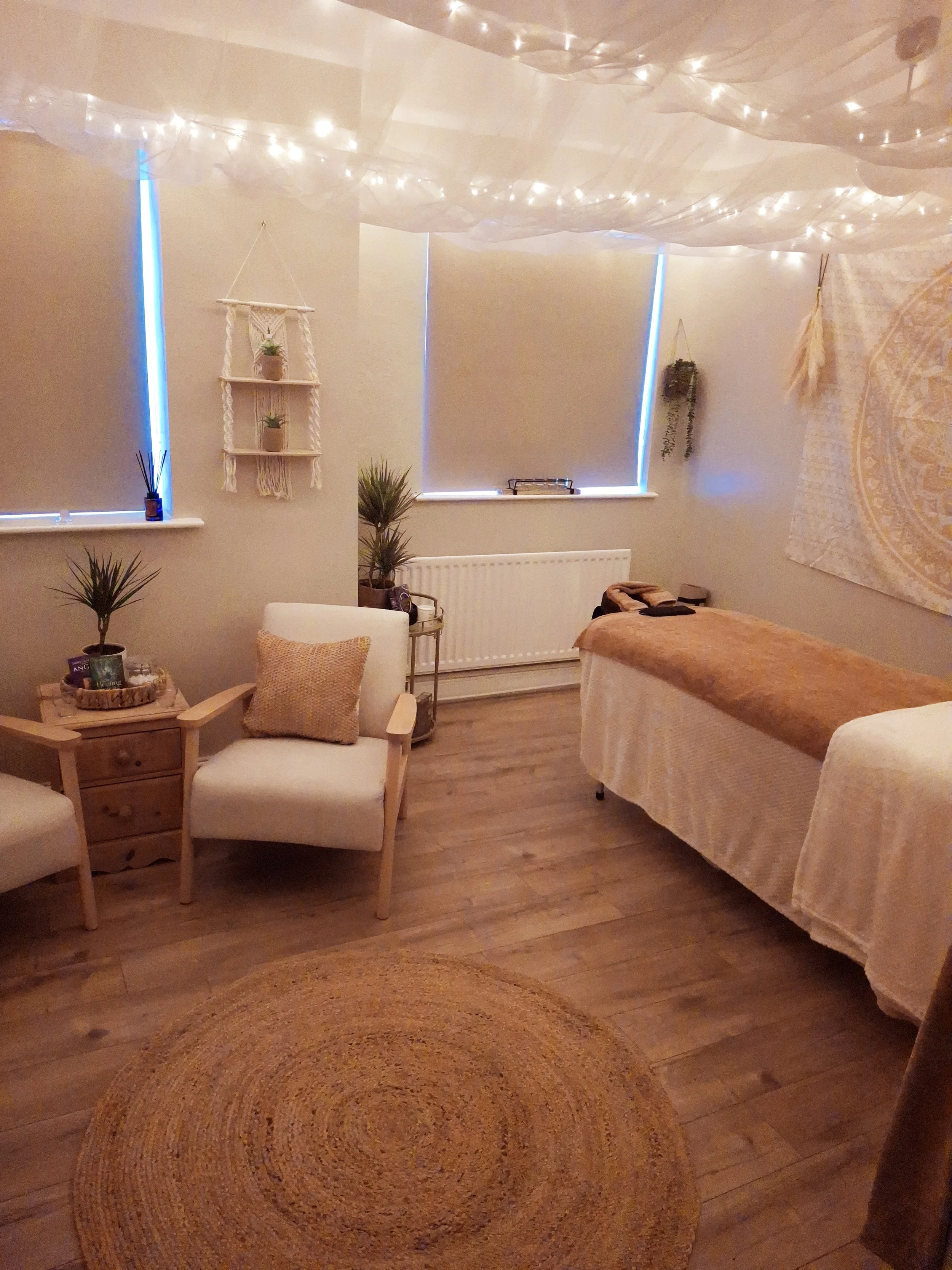 A cozy room decorated with string lights on the ceiling, a massage table covered with a beige blanket, a white armchair with a beige pillow, potted plants, a woven round rug, and window blinds.