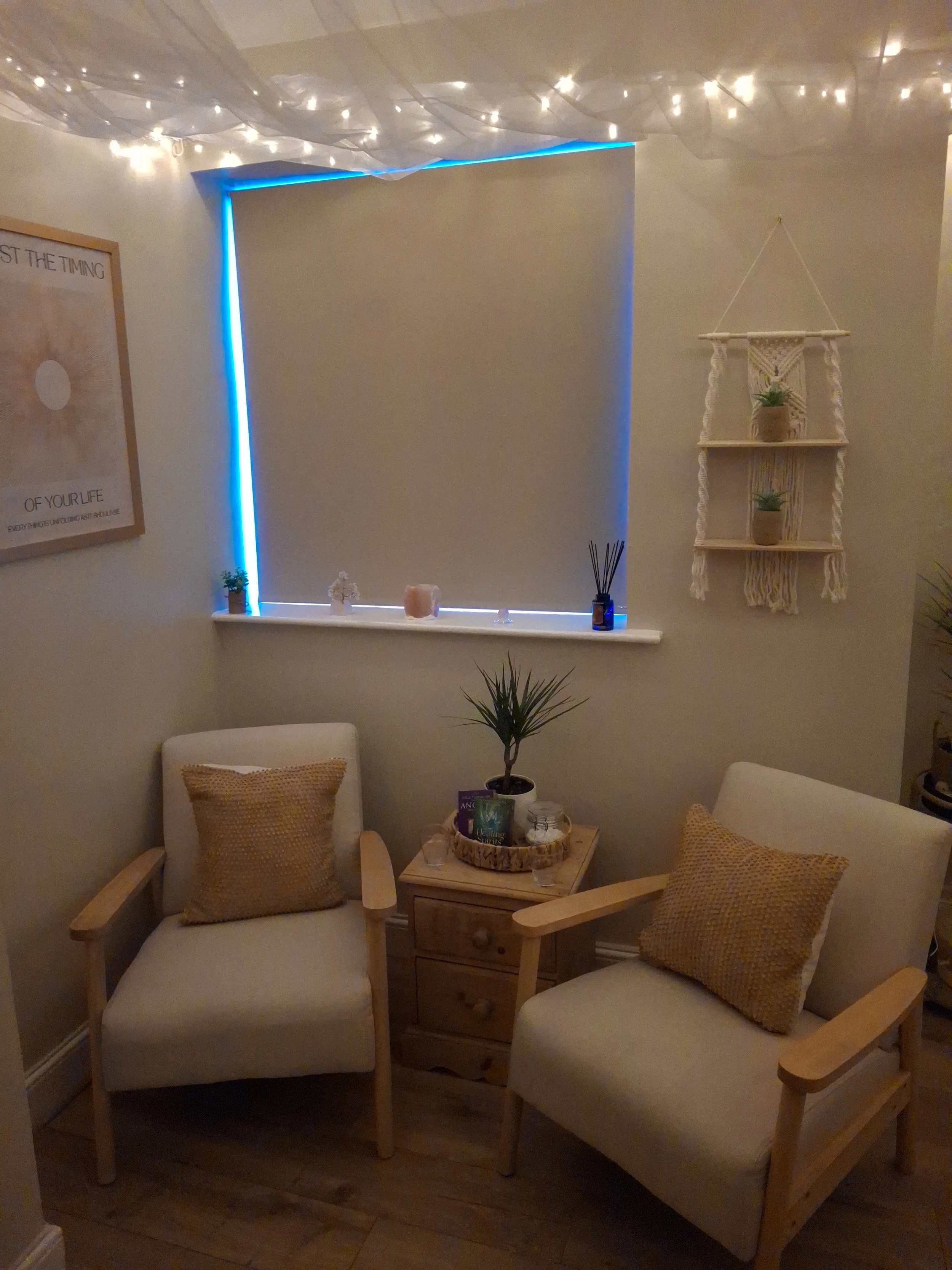 Cozy corner with two beige armchairs with yellow cushions, a small wooden side table with plants and candles, a window with blue LED strip lighting, string lights on the ceiling, and a macrame wall hanging with small plants.