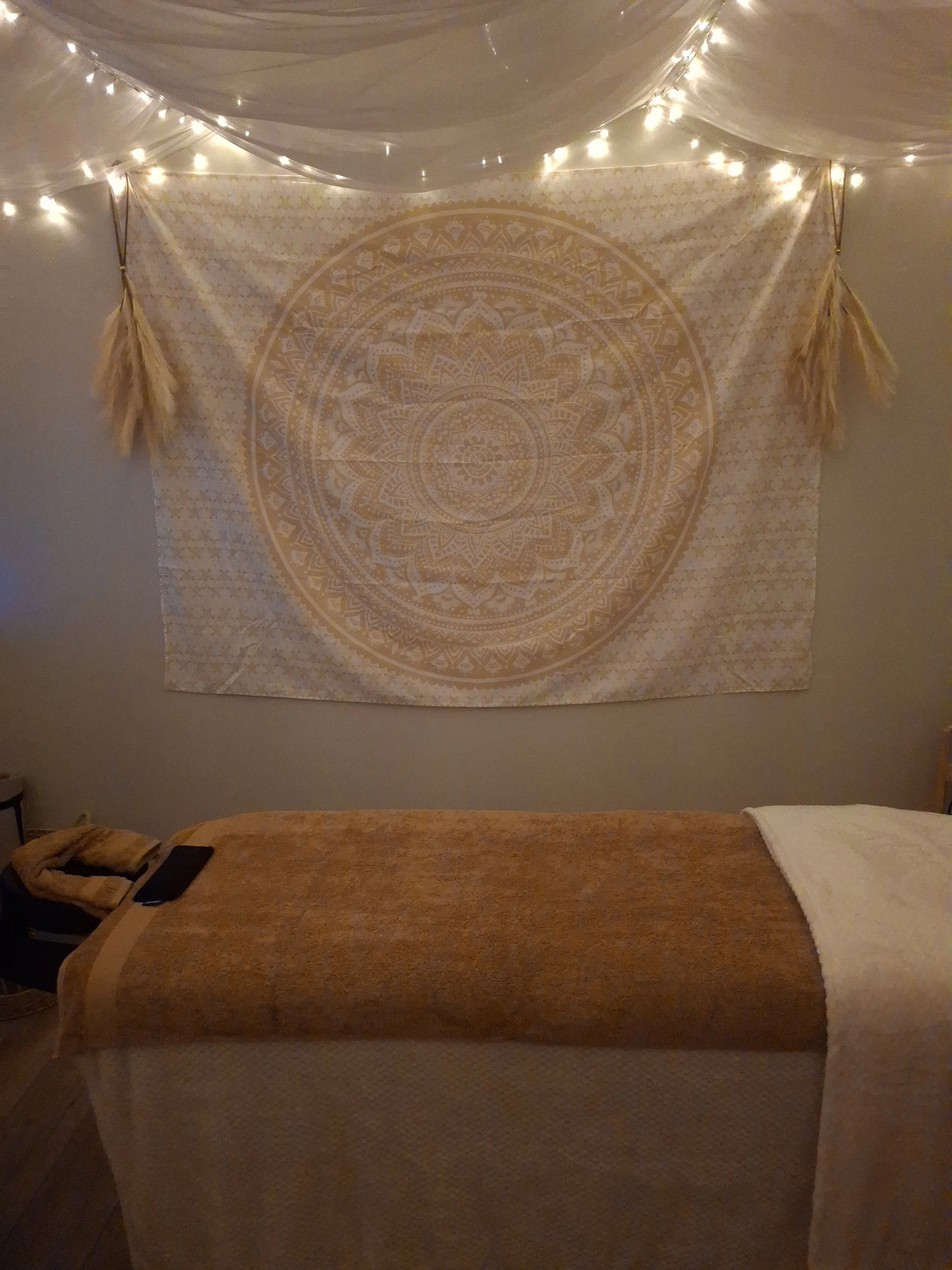 A decorated massage or therapy room with a massage table covered in a beige towel, a white blanket, and a tapestry with a mandala design hanging on the wall. String lights and hanging dried wheat decorations add warm ambient lighting.