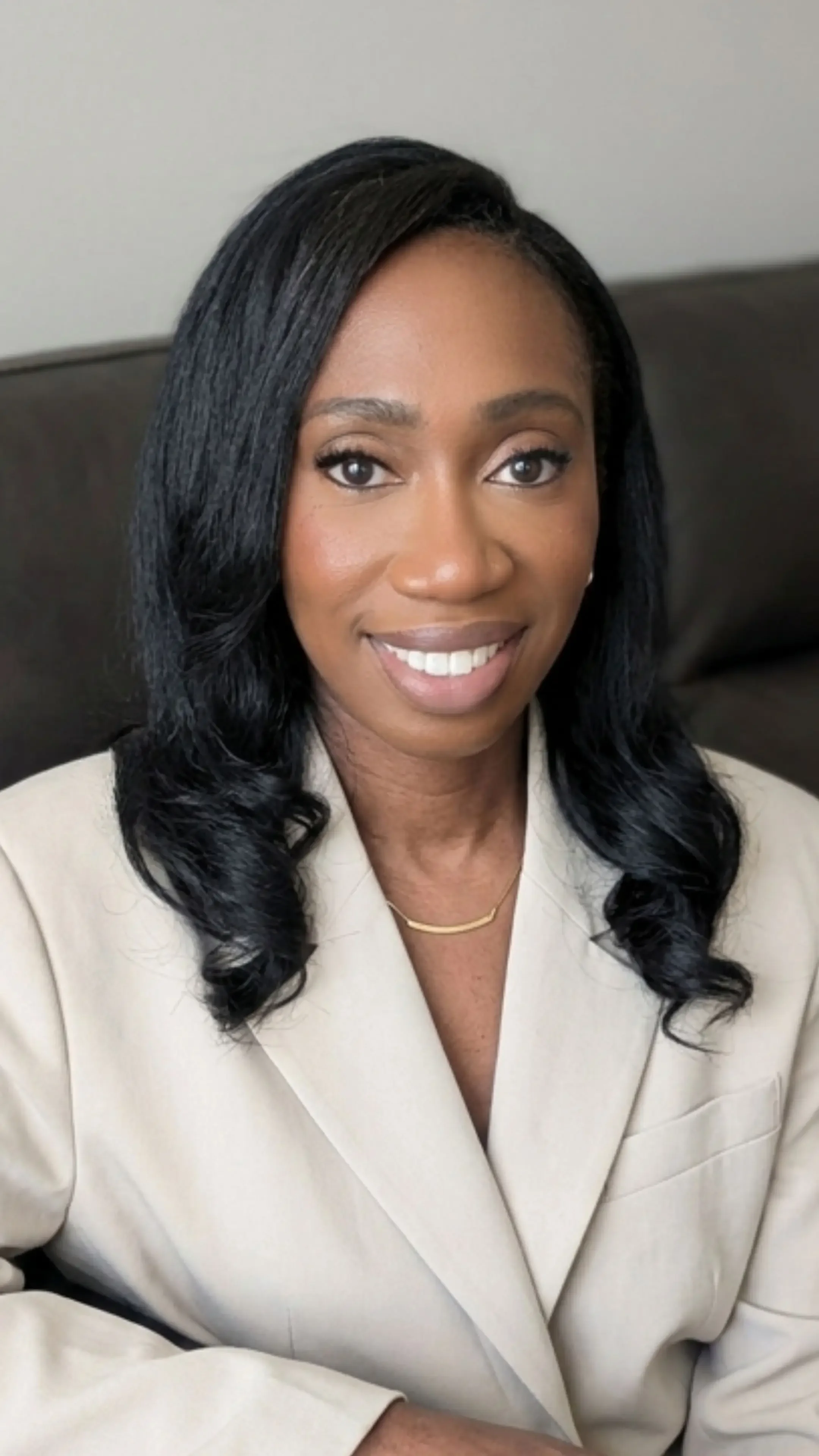 A smiling woman in a beige blazer, sitting and leaning on a dark leather couch.