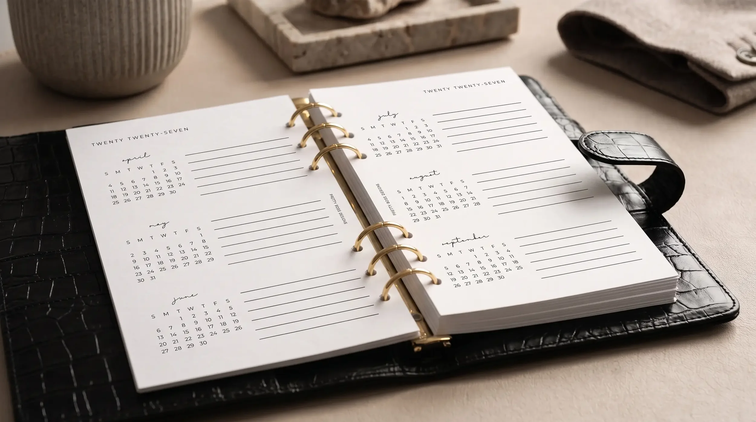 An open black crocodile-embossed leather ring binder displaying a 2027 year overview planner insert with small monthly calendars for April through September and blank lines beside each, styled on a beige surface with soft natural lighting.