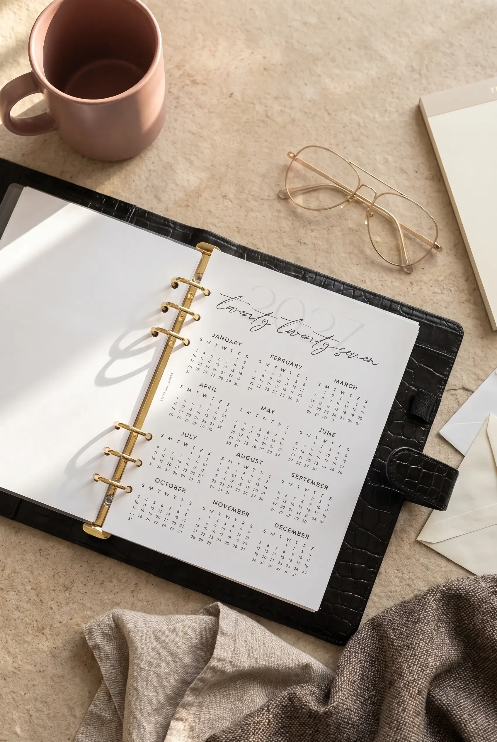 An open black leather A5 ring binder displaying a "twenty twenty-seven" year-at-a-glance calendar insert, styled on a textured beige surface with a pink mug, gold wire-rimmed glasses, envelopes, and a notebook.
