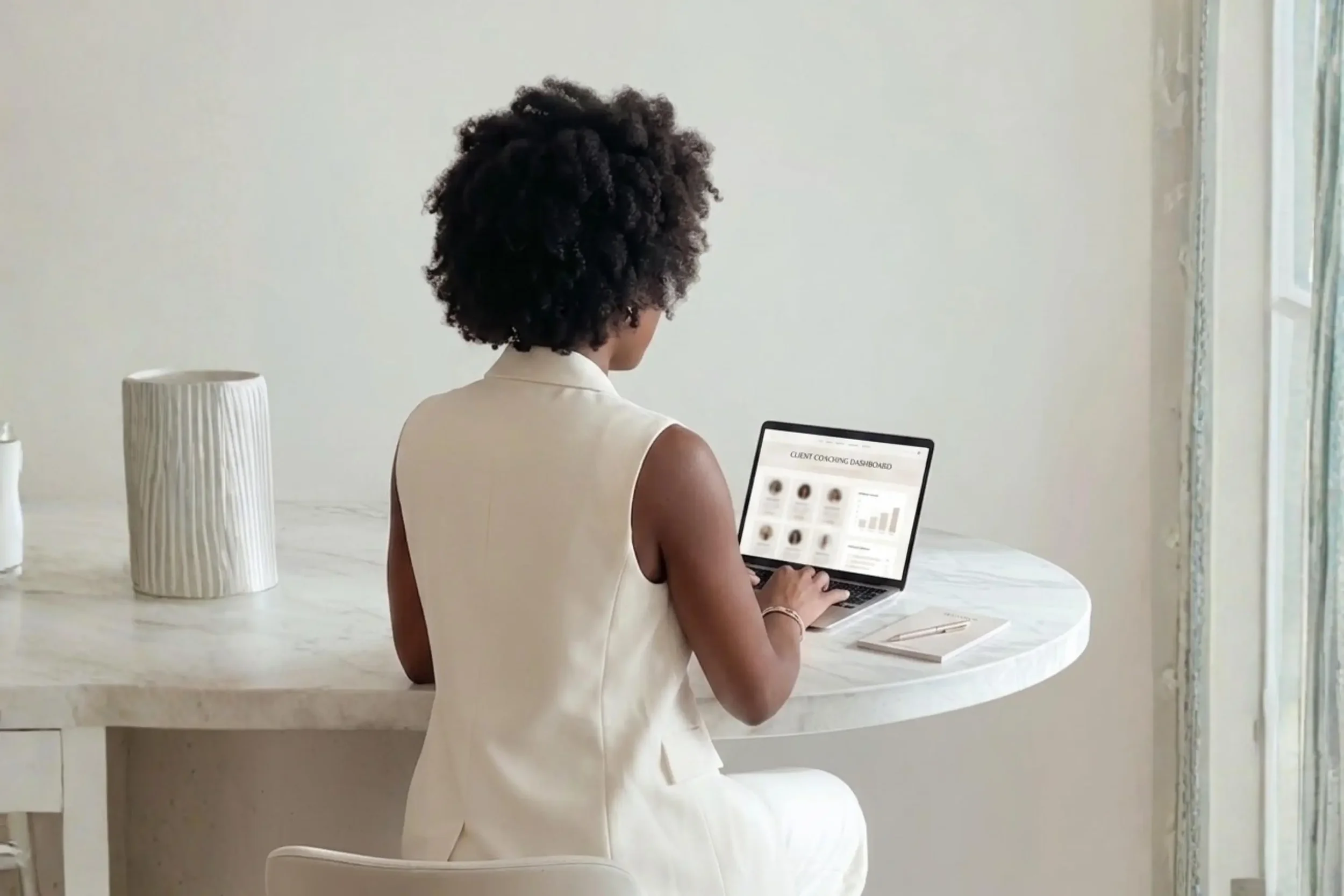 Back view of a woman with curly hair sitting at a marble table and typing on a laptop.
