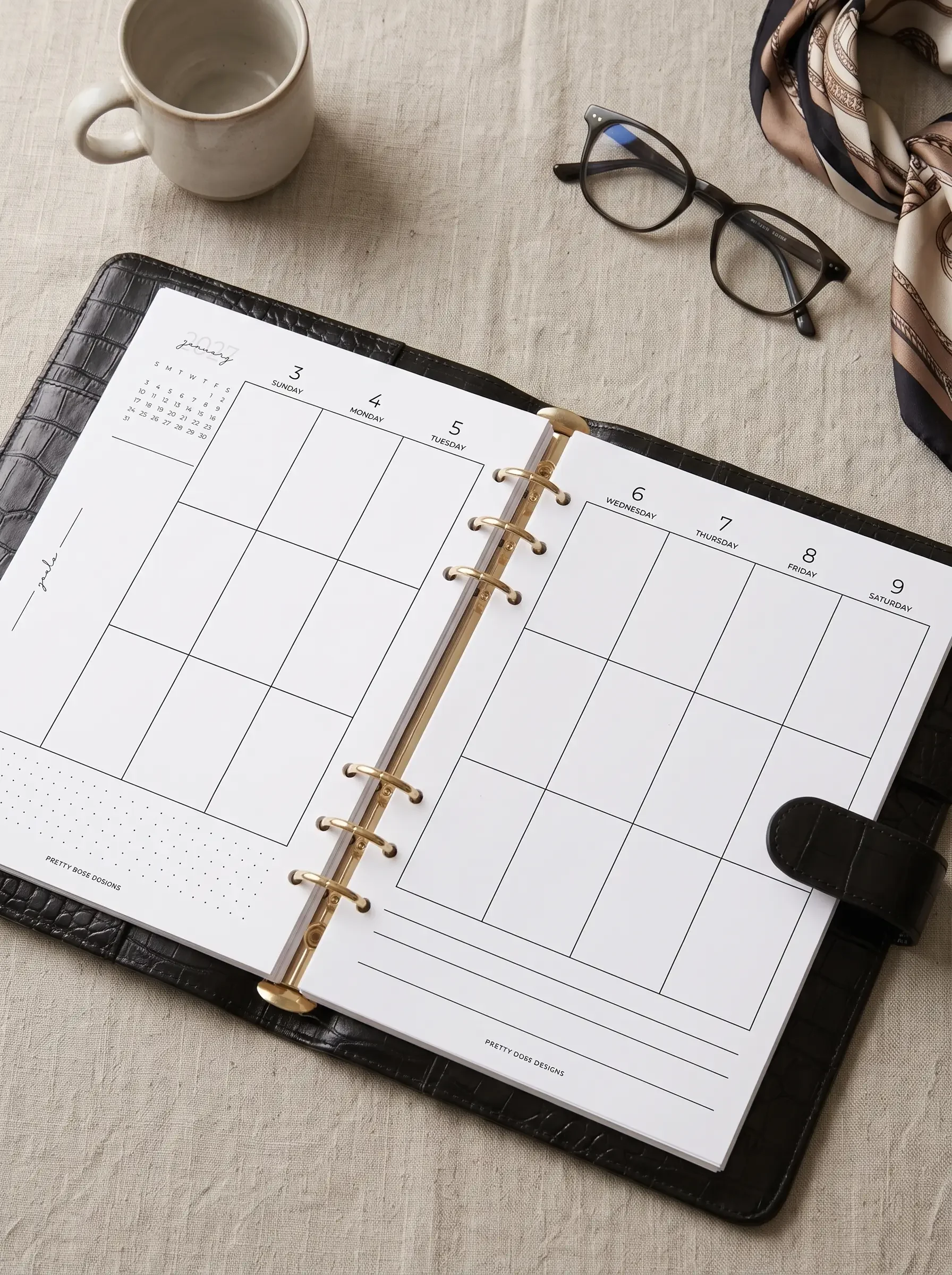 An open black leather A5 ring binder featuring a blank vertical weekly planner spread with a goals section, arranged on a textured surface alongside a ceramic mug, dark-rimmed glasses, and a patterned silk scarf.