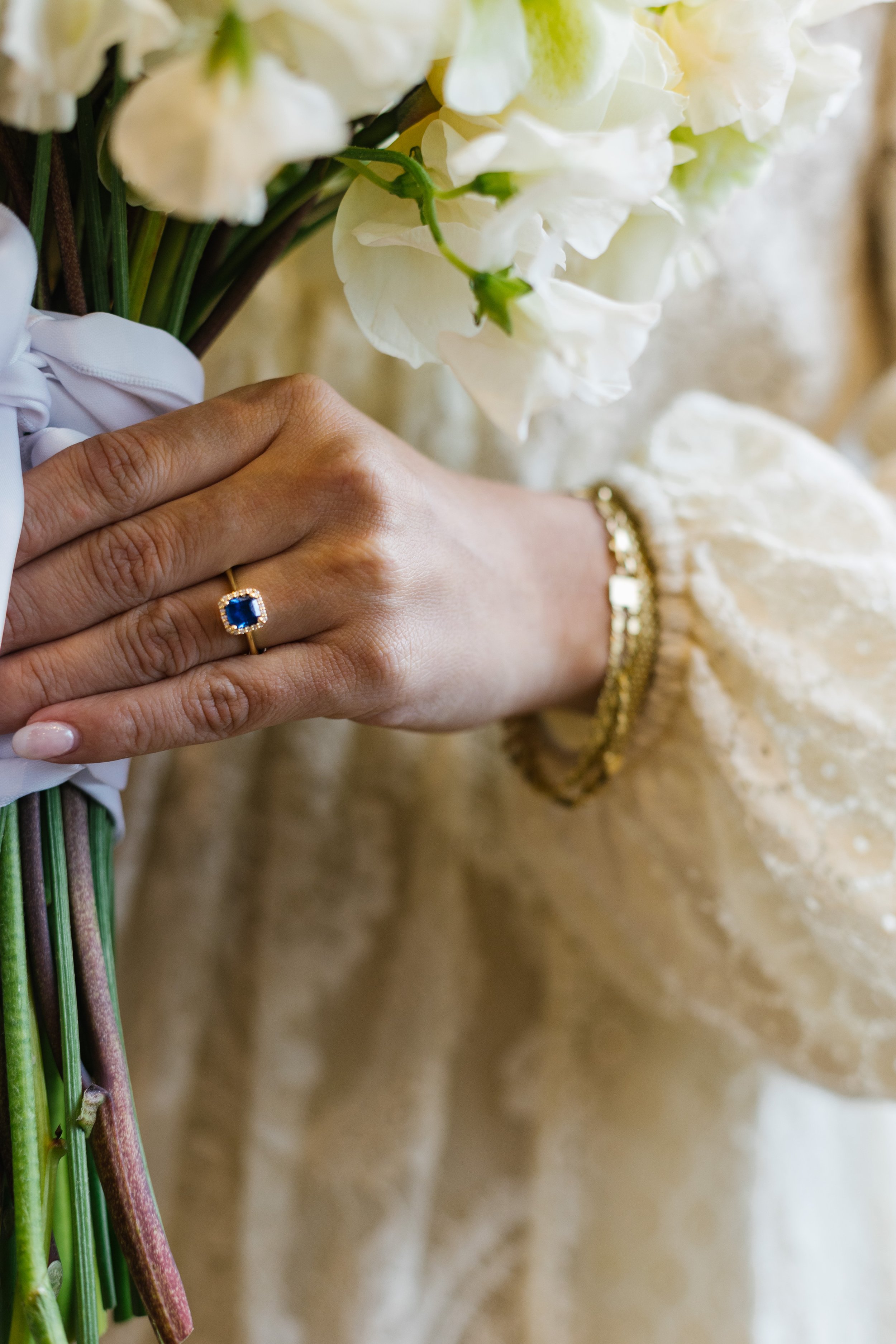 Close-up of a woman's hand resting on a wedding dress, showing a gold ring with a blue gemstone, gold bracelet, and holding a bouquet of white flowers