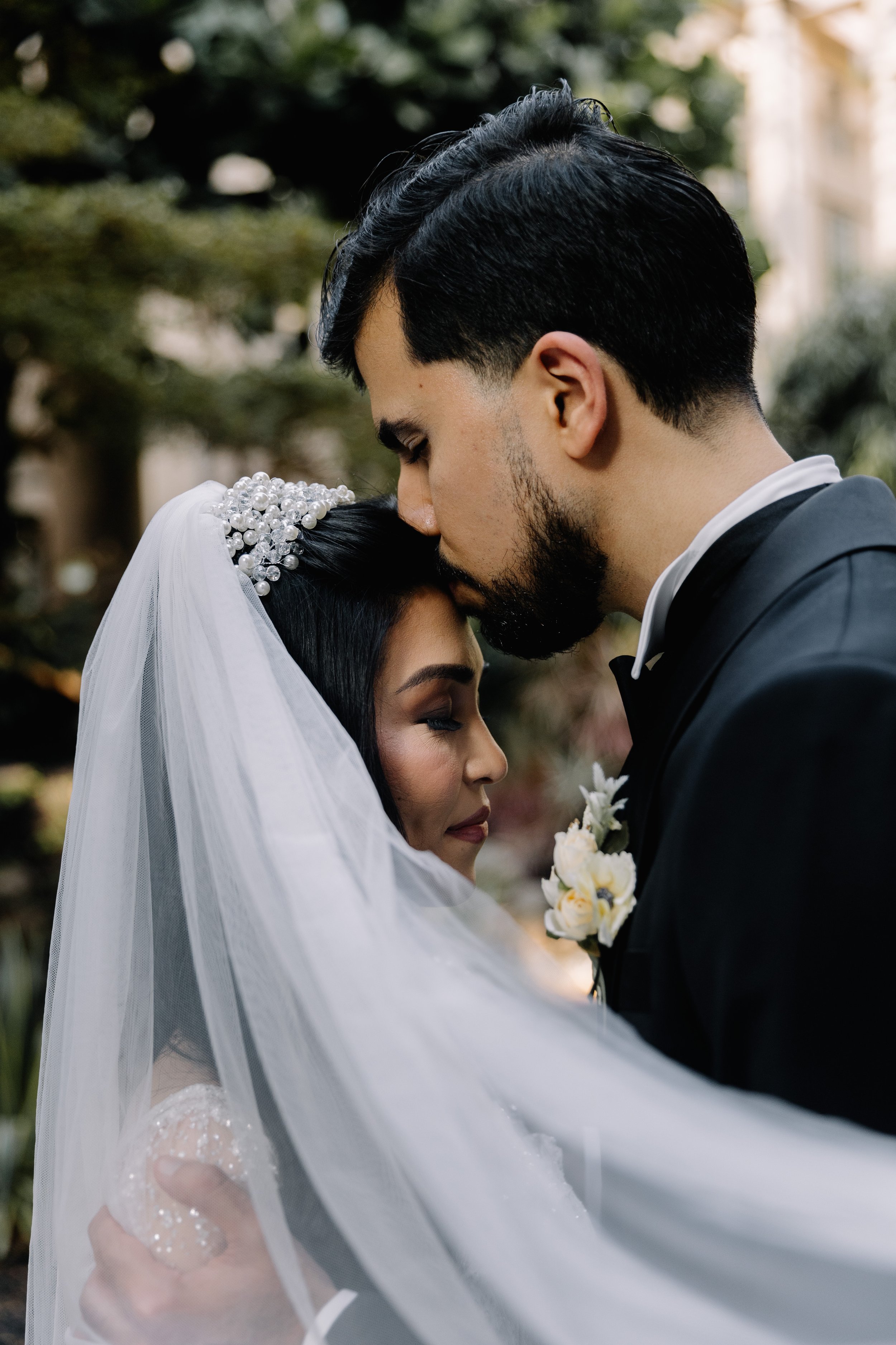 A happy bride and groom embrace outdoors, with the groom kissing the bride's forehead, surrounded by greenery.