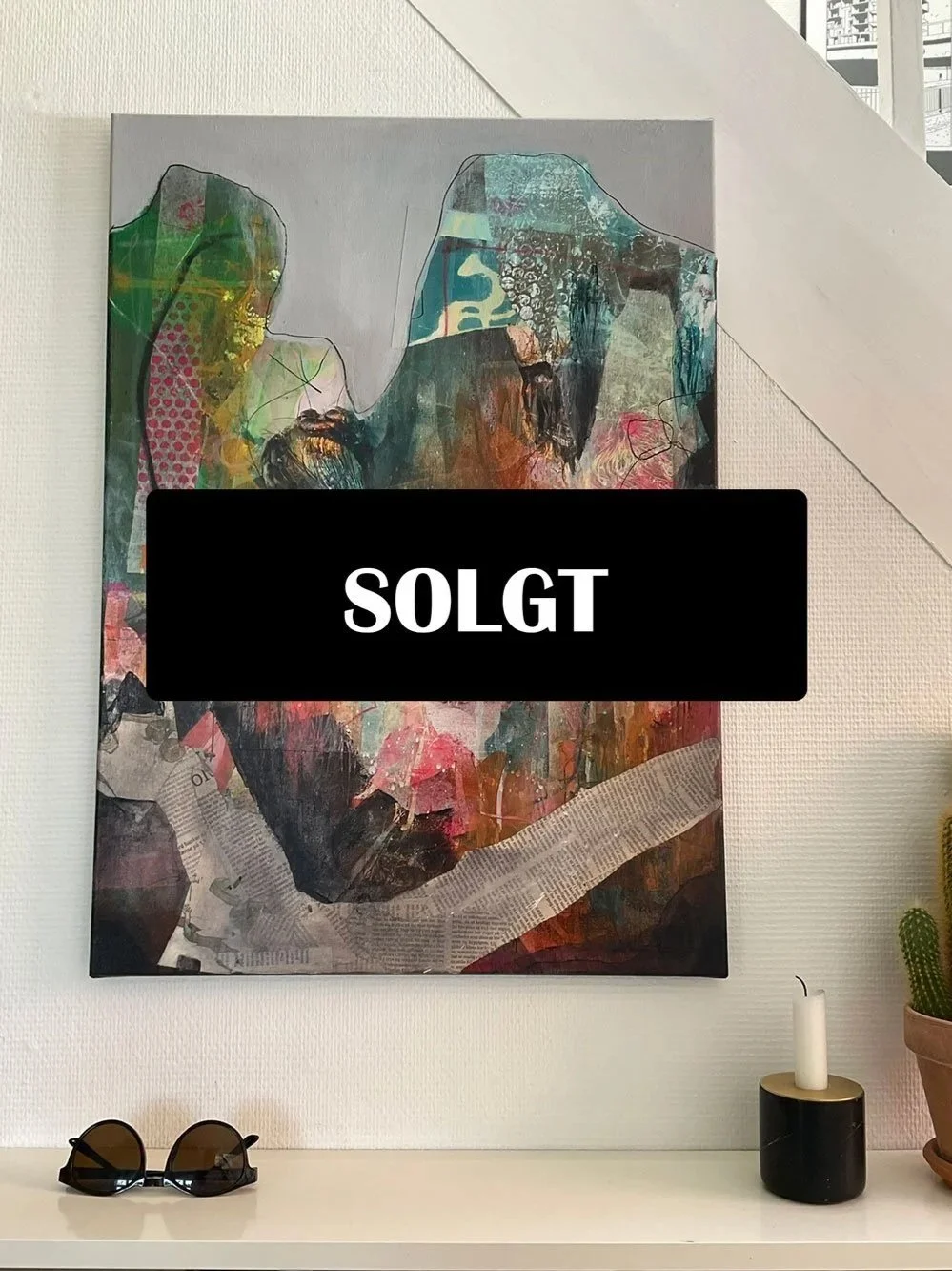 Moderne kunstværk med abstrakte elementer og en portrætfotografi af en kvinde med solbriller, hængende på en hvid væg over en hvid skænk.