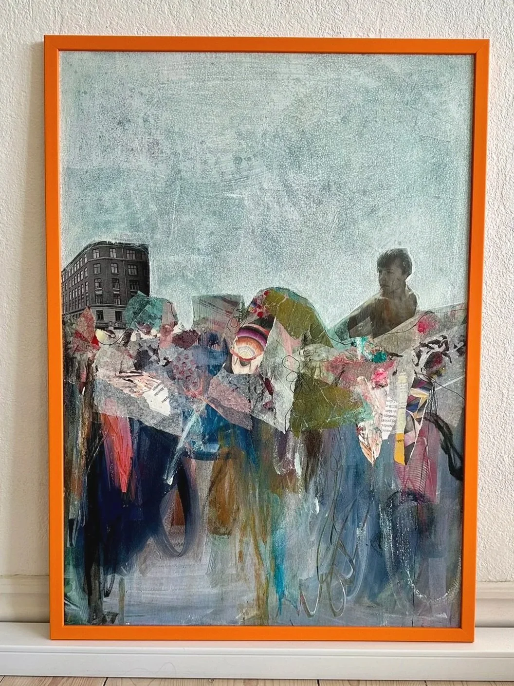 Abstrakt kunstværk med farverige papirklip, malet over en grå baggrund. Der er en lille del af en bygning i venstre side og en person med mørkt hår til højre.