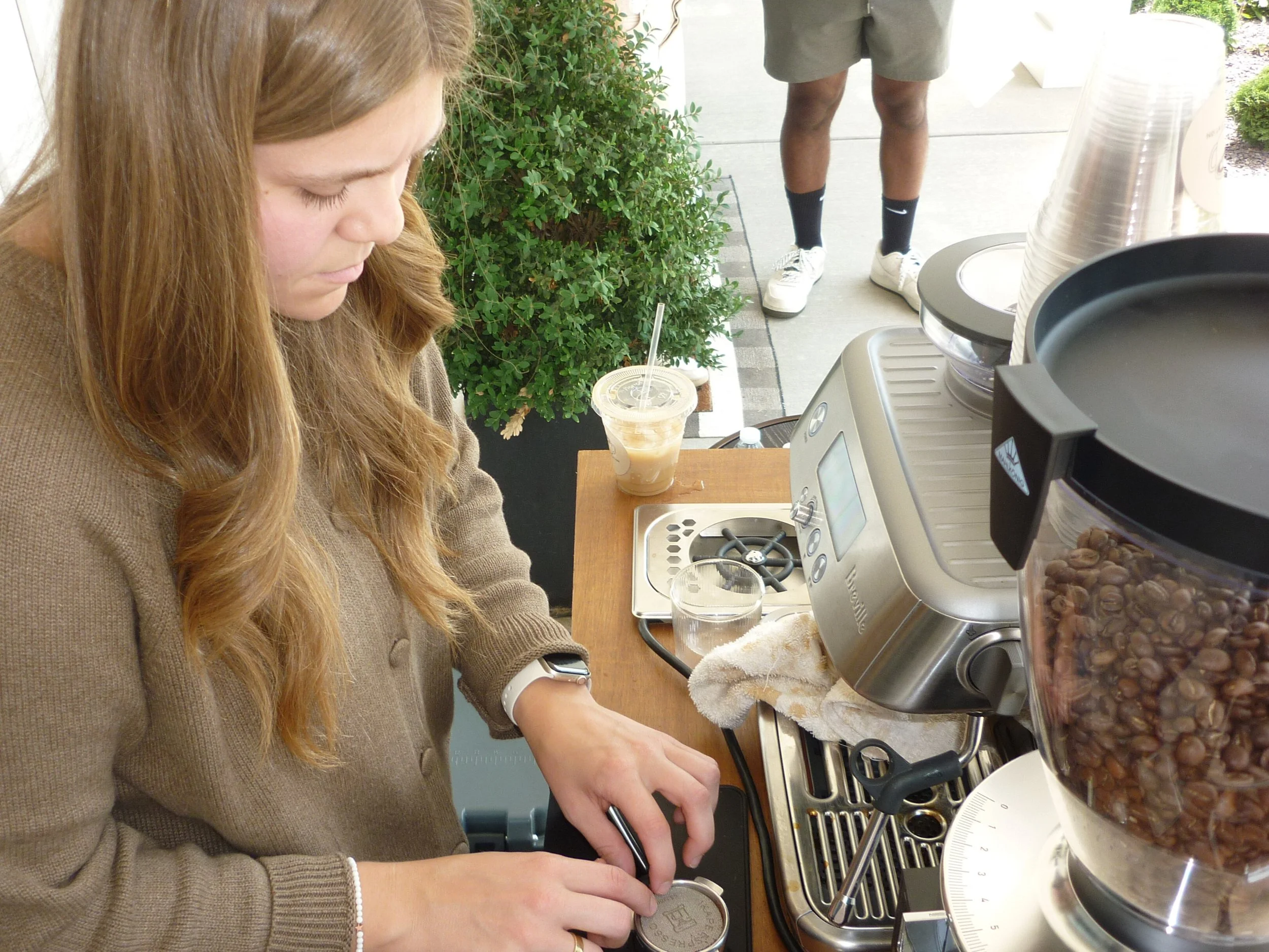 A woman making coffee at a coffee shop counter, with a coffee grinder and coffee beans visible.