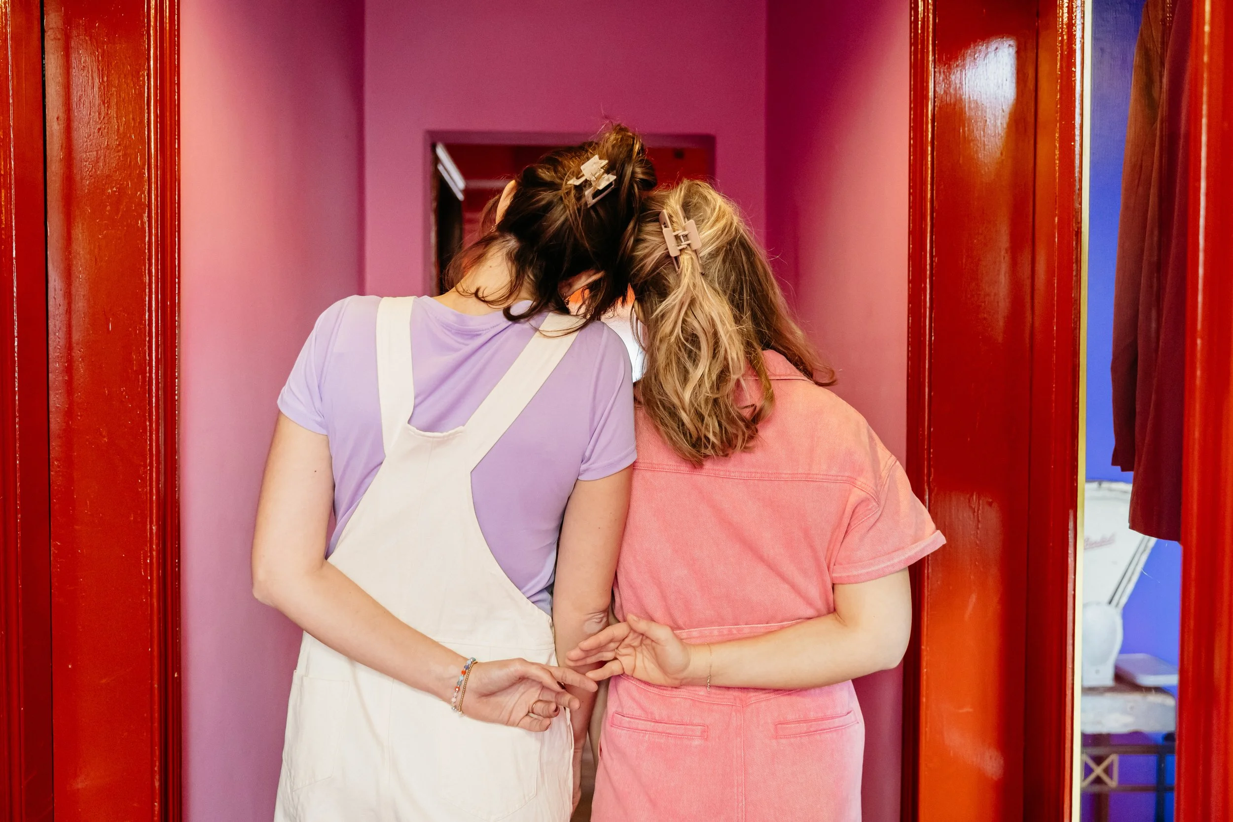 Twee jonge vrouwen met hun hoofden dicht tegen elkaar, in een kleine ruimte met roze en blauwe muren, en houten deurkozijnen. Ze dragen haarbandjes en ontspannen kleding.