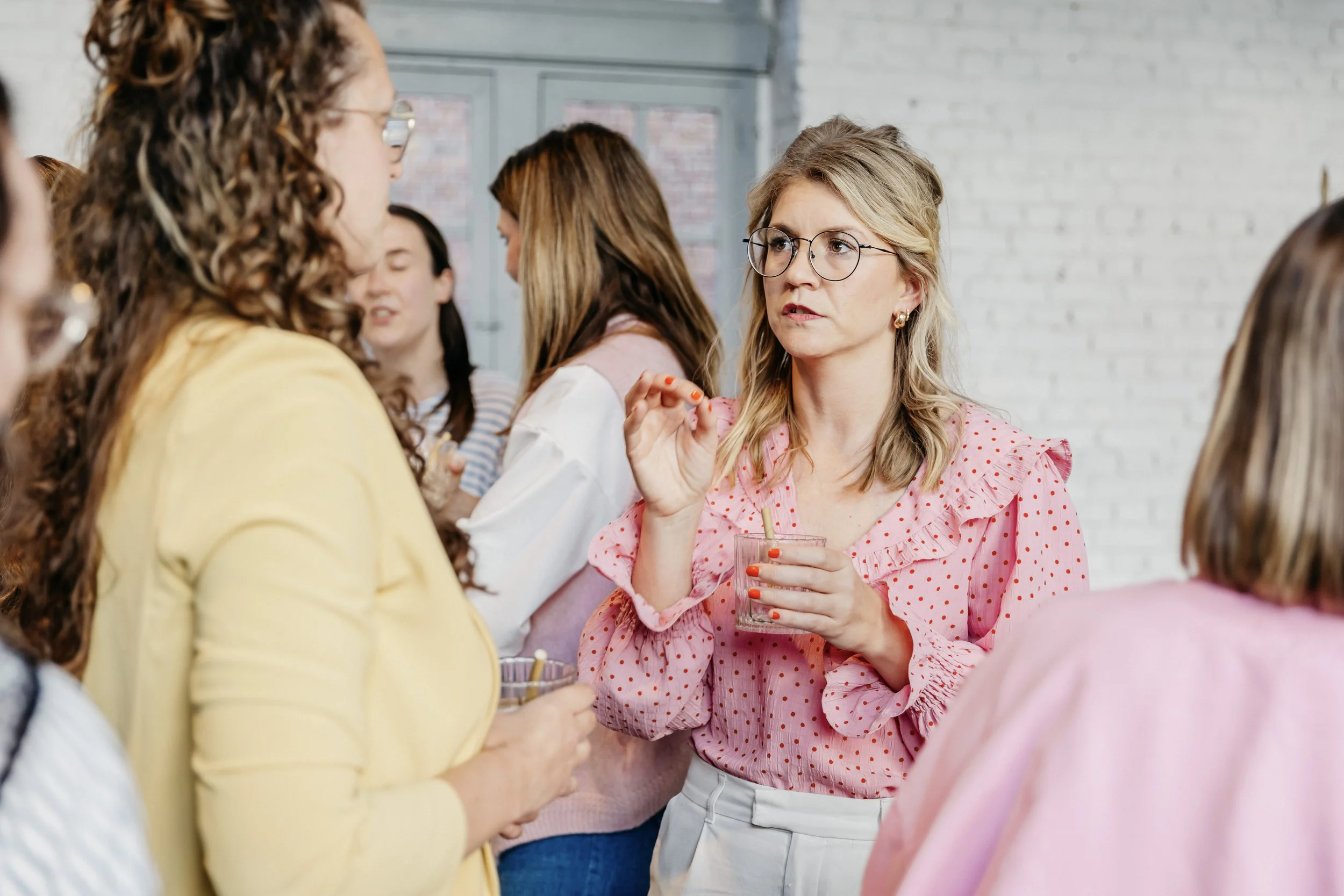 Kim met blond, golvend haar, glazen en een roze blouse met rode stippen, die een drankje vasthoudt en praat met een vrouw in een gele jas, op een sociale bijeenkomst van Kop op compagnie.