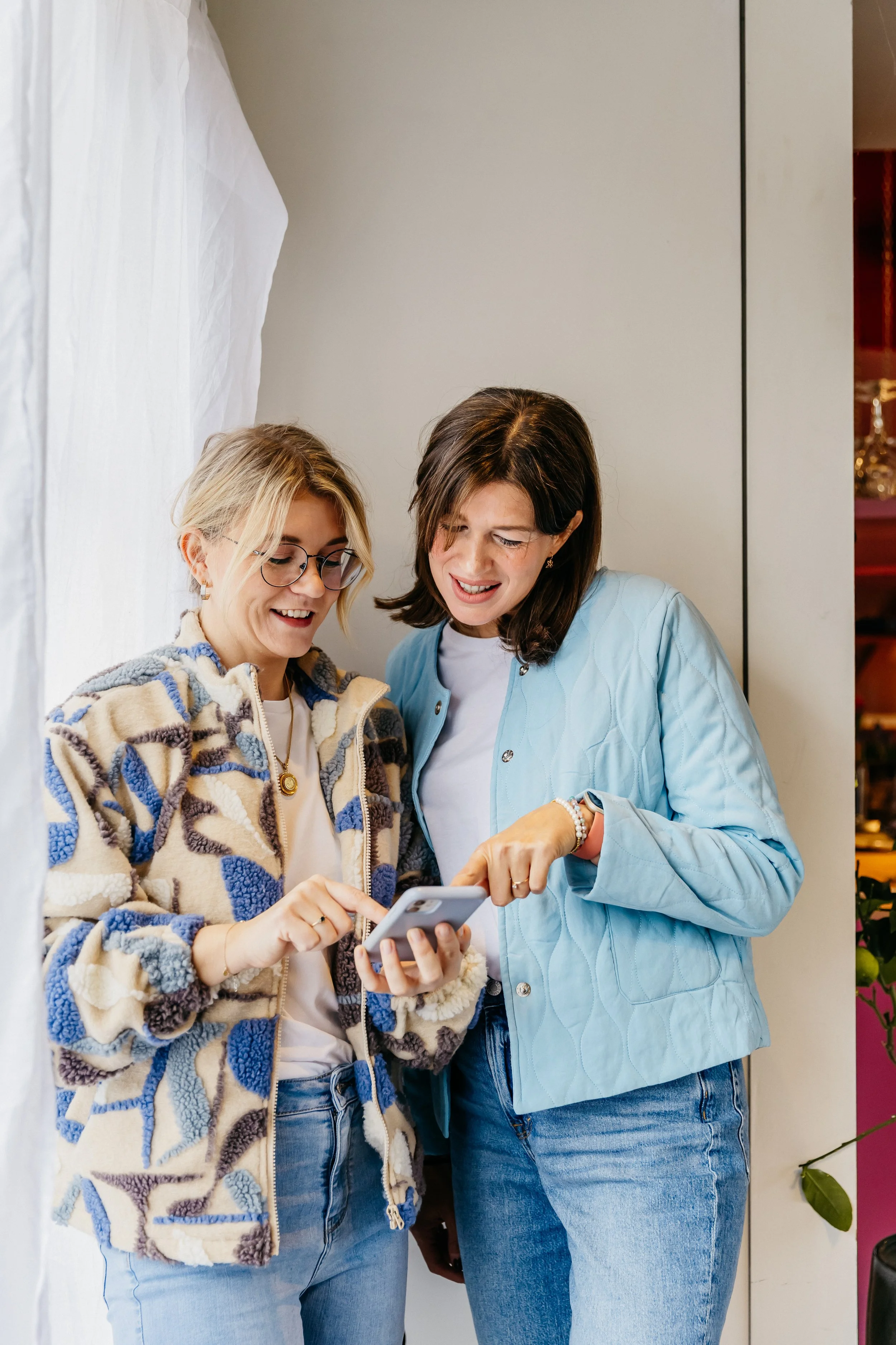 Twee vrouwen, Kim en Sofie, kijken samen naar een smartphone en lachen, ze staan binnen bij een muur.