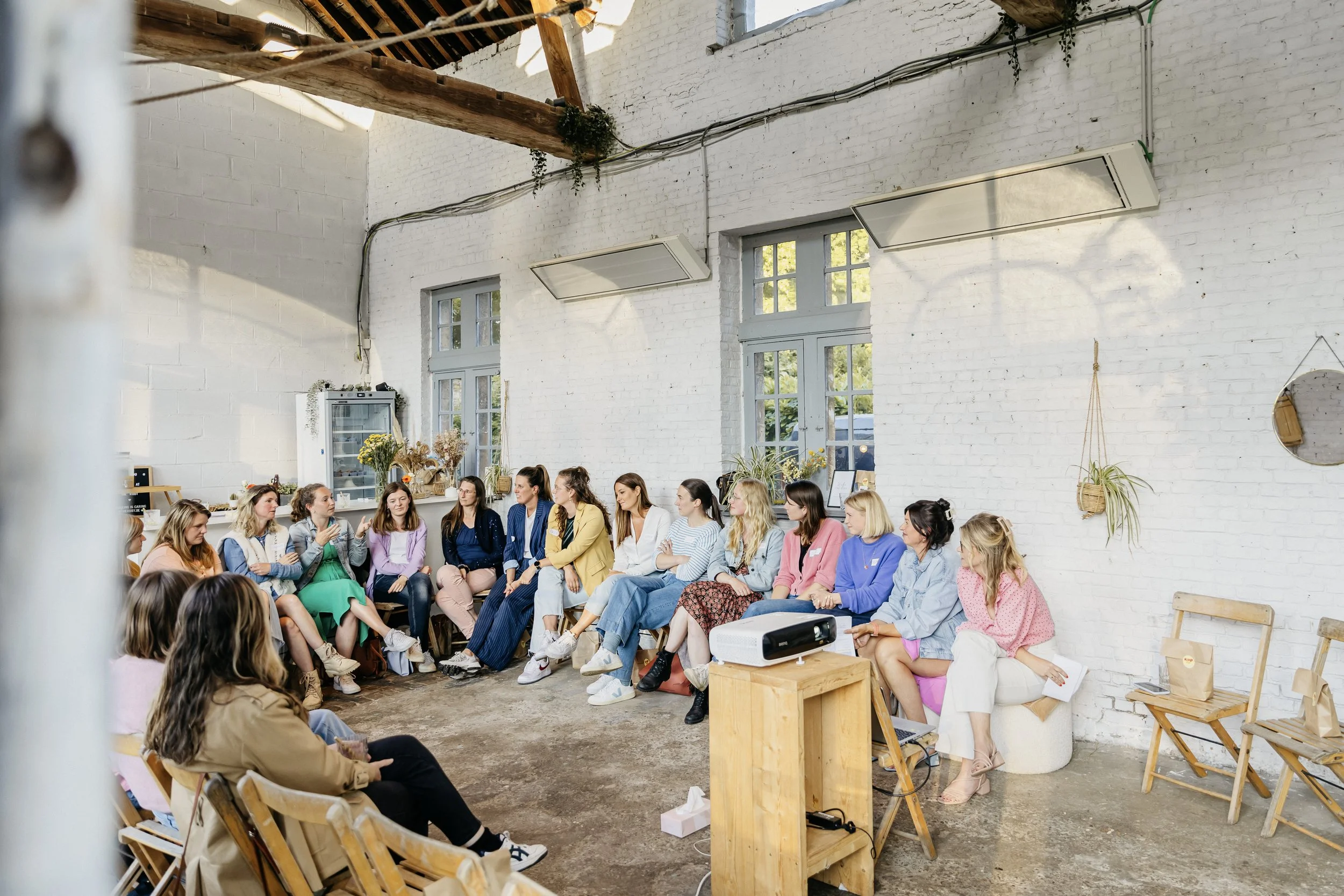 Een groep vrouwen zit in een ronde in een lichte zaal met witte muren en grote ramen, mogelijk tijdens een workshop of bijeenkomst van Kop op Compagnie.