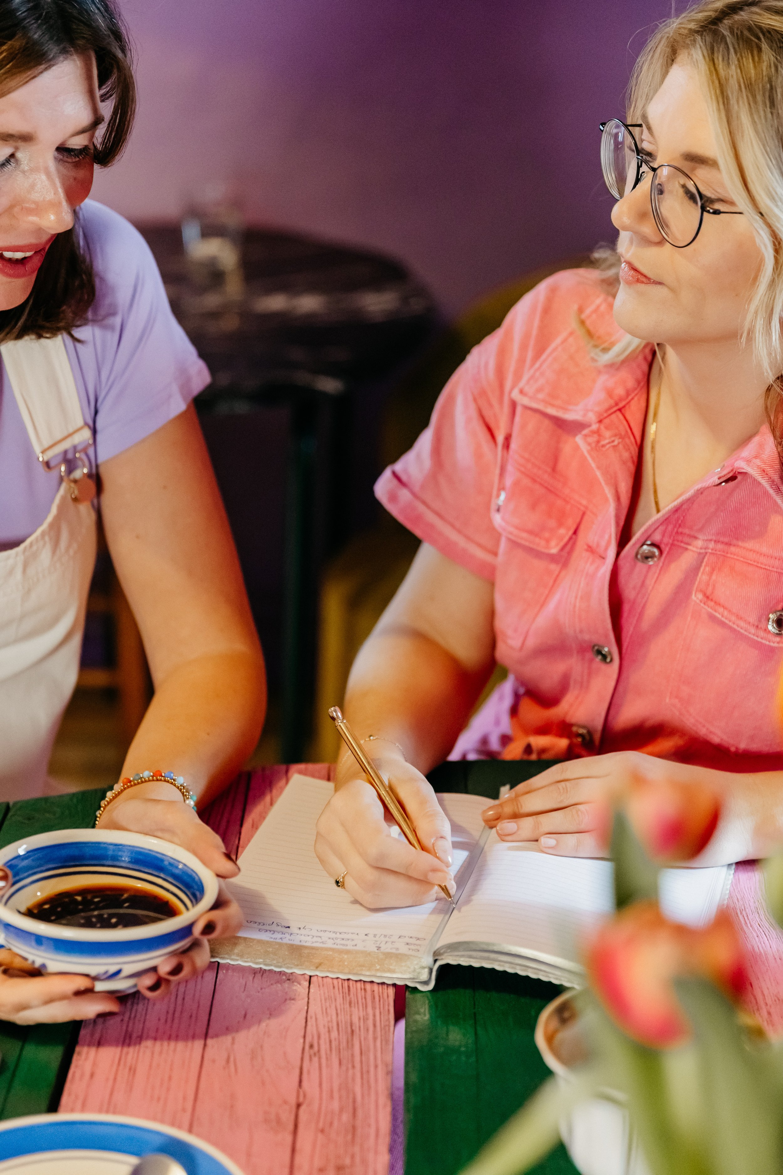 Twee vrouwen zitten aan een kleurrijke eettafel, de ene schrijft in een notitieboekje, de andere houdt een kom met een koffie vast.