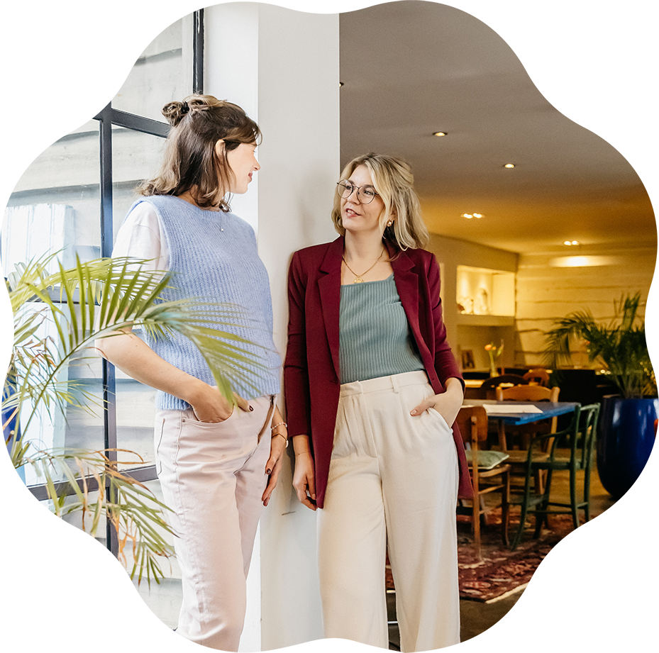 Twee vrouwen praten in een interieur met grote ramen, planten en een eettafel in de achtergrond.
