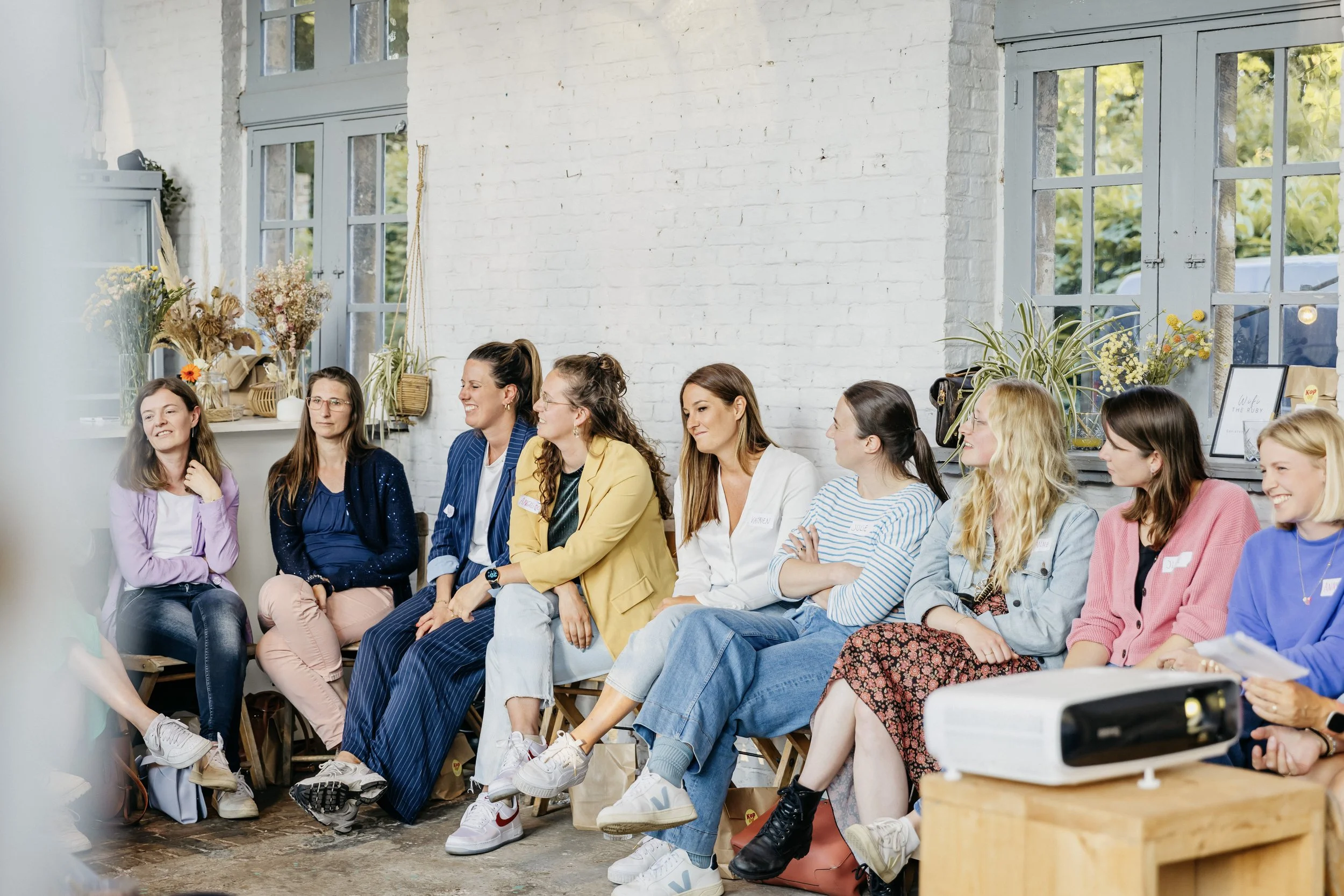 Een groep vrouwen zit op een bank in een lichte, gezellige kamer met grote ramen, decoraties en planten, tijdens een bijeenkomst of workshop van Kop Op Compagnie.