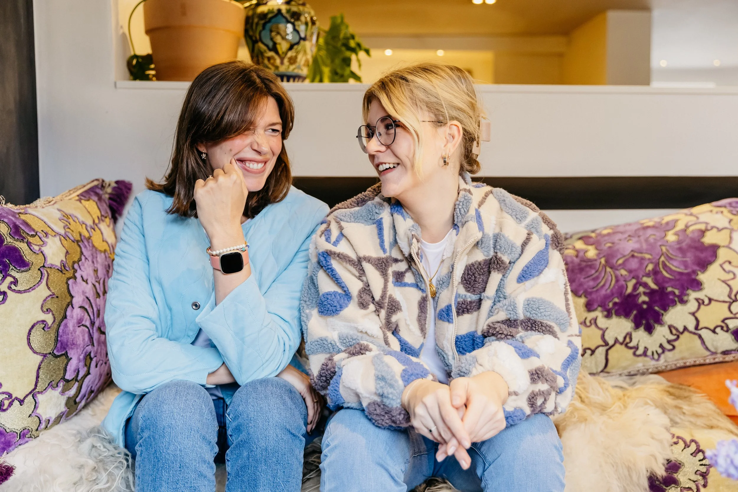 Twee vrouwen die samen lachen op een bank met kleurrijke kussens.