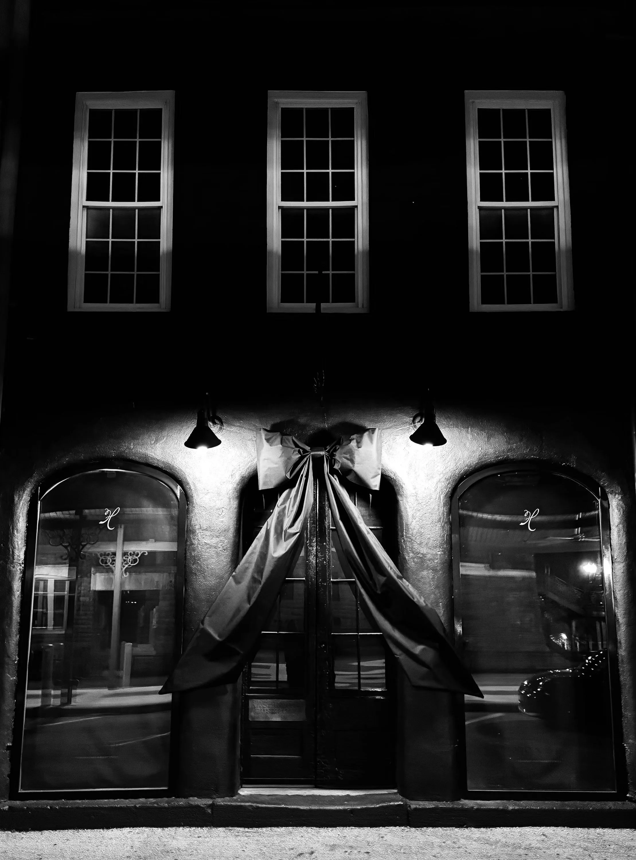 Black and white photo of a storefront with large glass windows, a door with glass panes, a large bow on the door, and two lights above the door. Four windows are visible above.